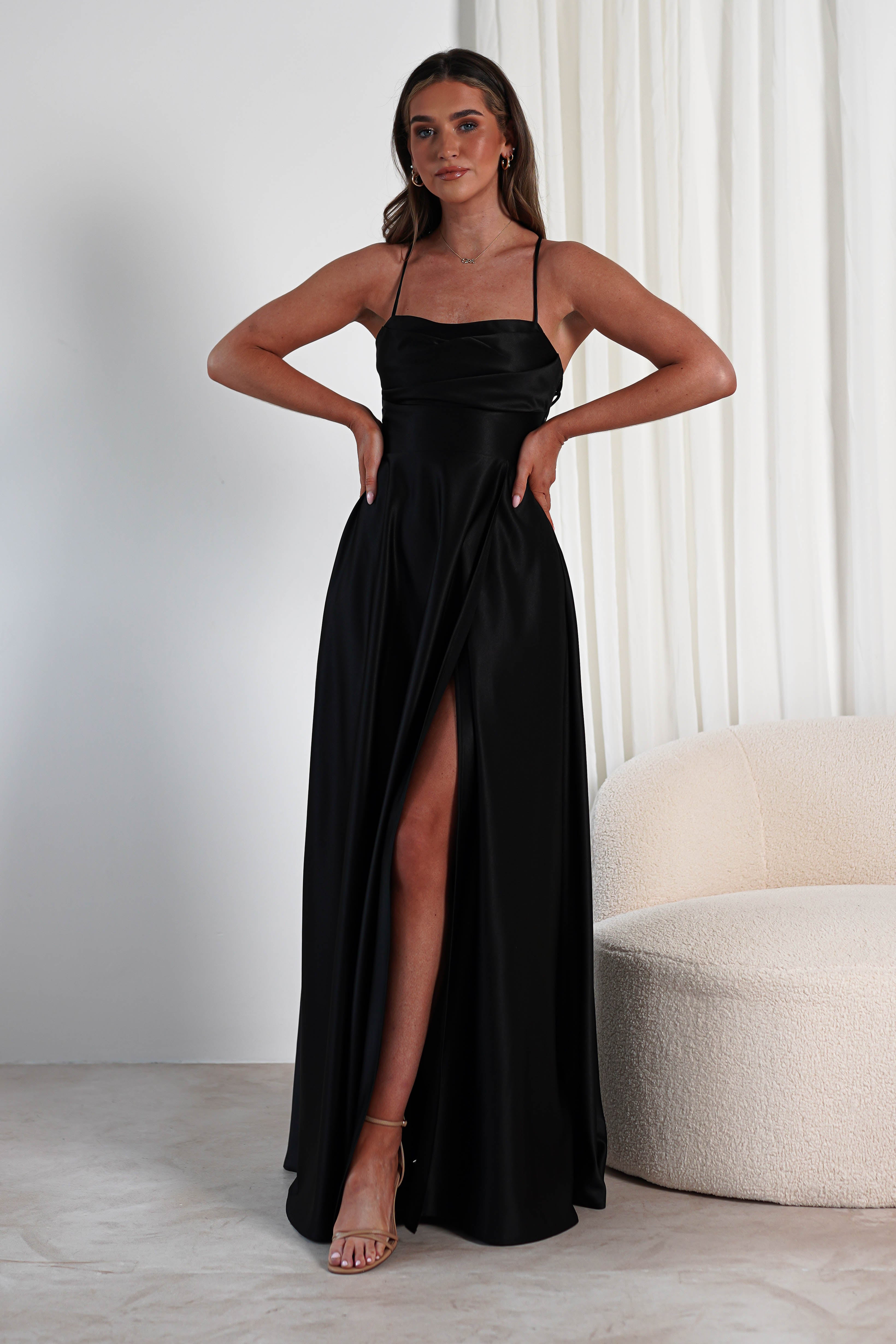 A woman stands confidently in a sleek, black satin dress featuring a cowl neckline and high slit. She poses with hands on hips near a white, modern chair and curtain. The room embraces a minimalist style with a light, neutral color palette.