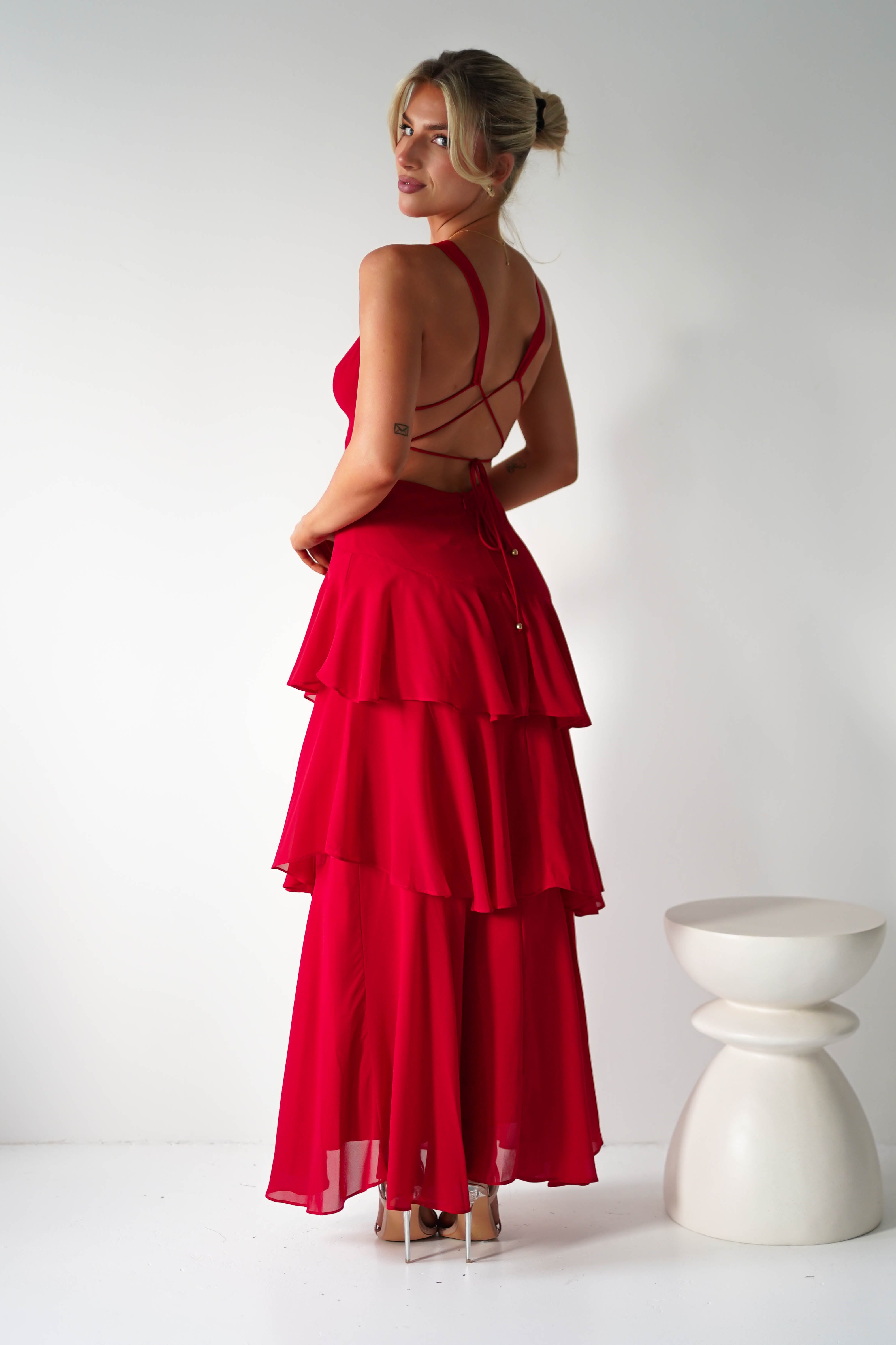 Woman in a red, tiered chiffon maxi dress stands sideways against a white background. Her blonde hair is styled in a bun, and the cross-strap back perfectly complements her chic wedding guest attire. A small white stool is placed to her left.
