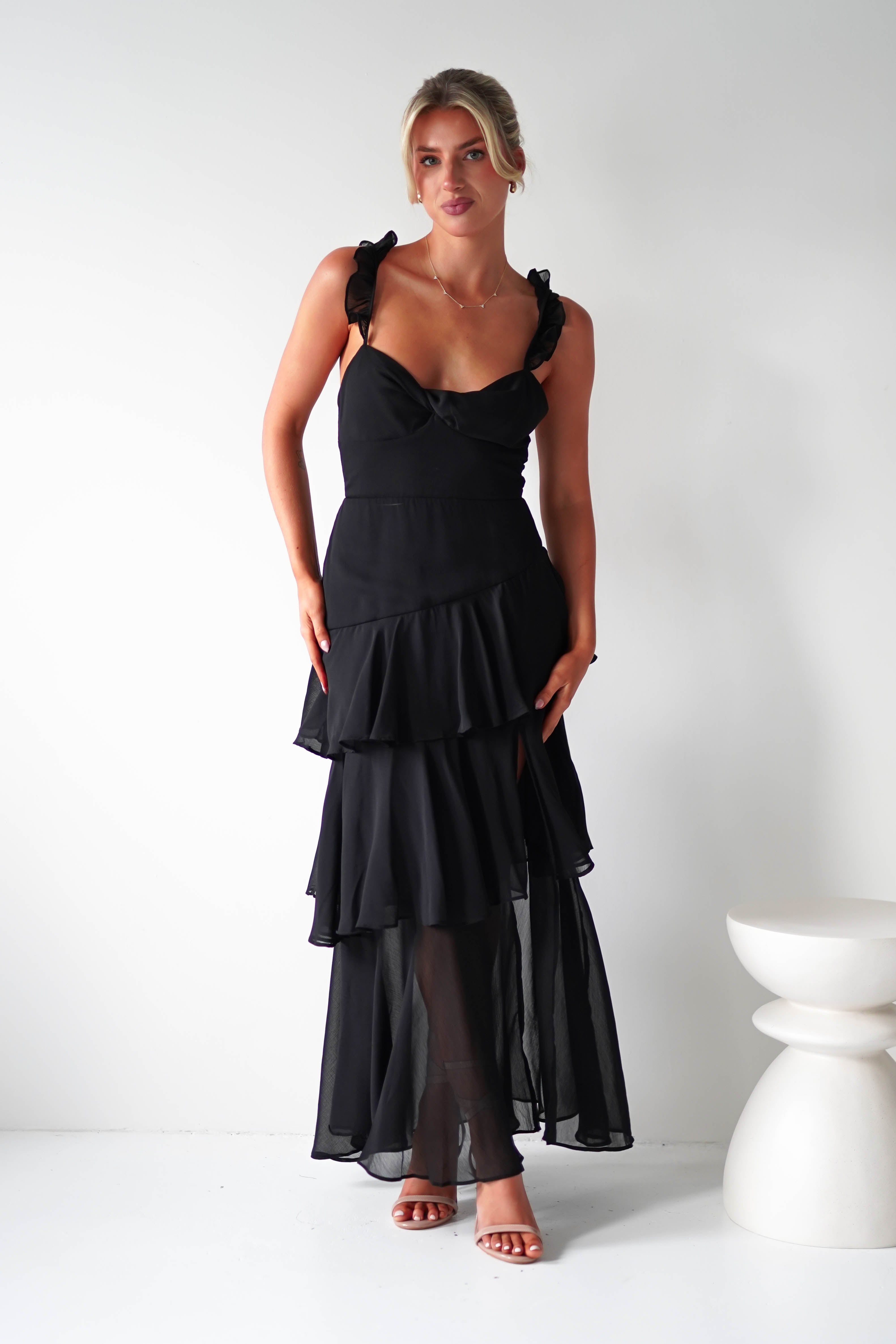 A woman in a tiered black ruffle dress stands against a plain white wall. The dress, crafted from chiffon fabric, features ruffled straps and a semi-sheer lower section. With strappy heels and her hair styled up, she exudes elegance, perfect for any wedding guest. A white decorative stool is visible in the background.