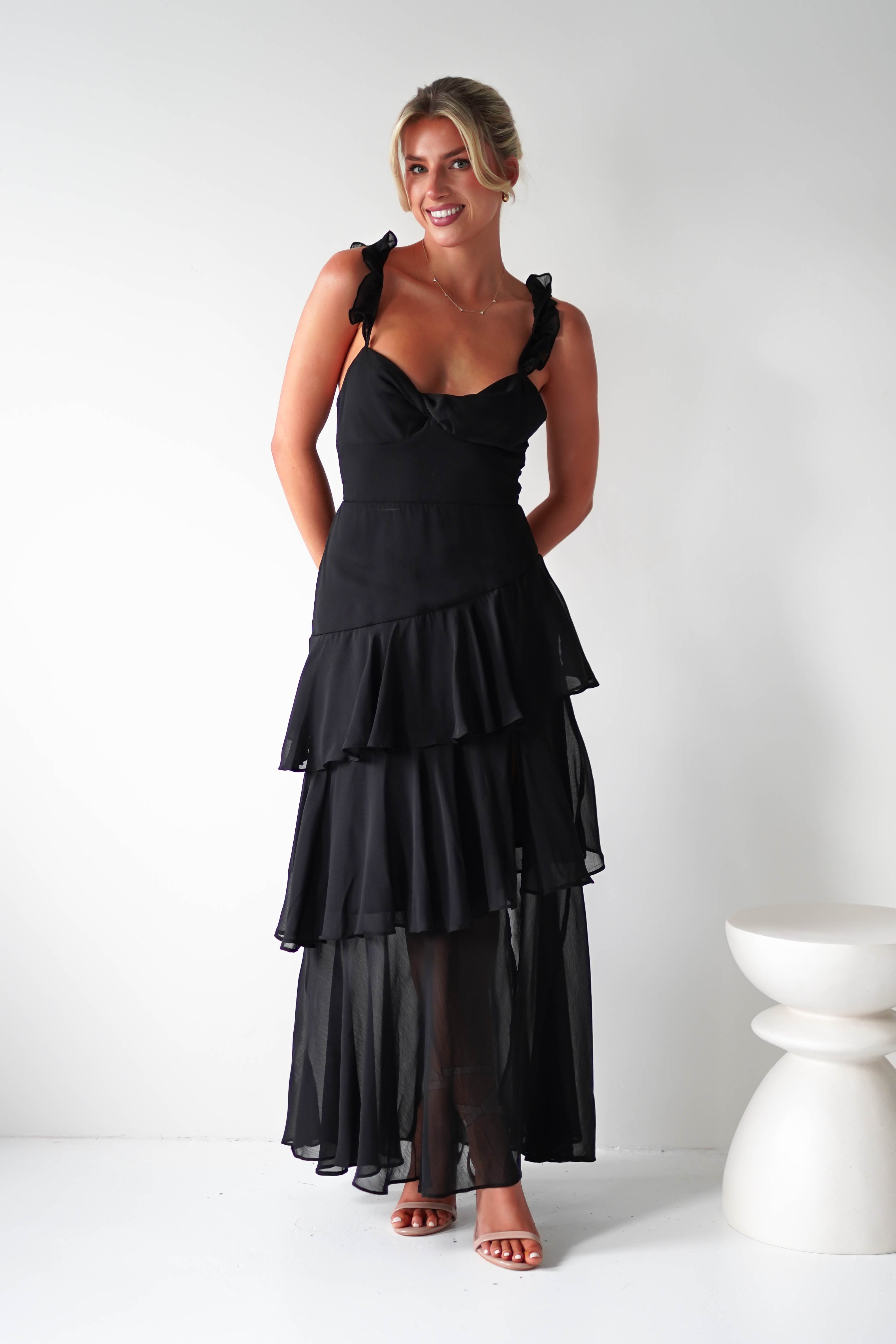 Woman in a black chiffon tiered spaghetti strap dress and sandals, standing against a plain white background. She is smiling and looking slightly to the side, with hands behind her back, as if ready for a joyful occasion. A small white stool is visible on the right.