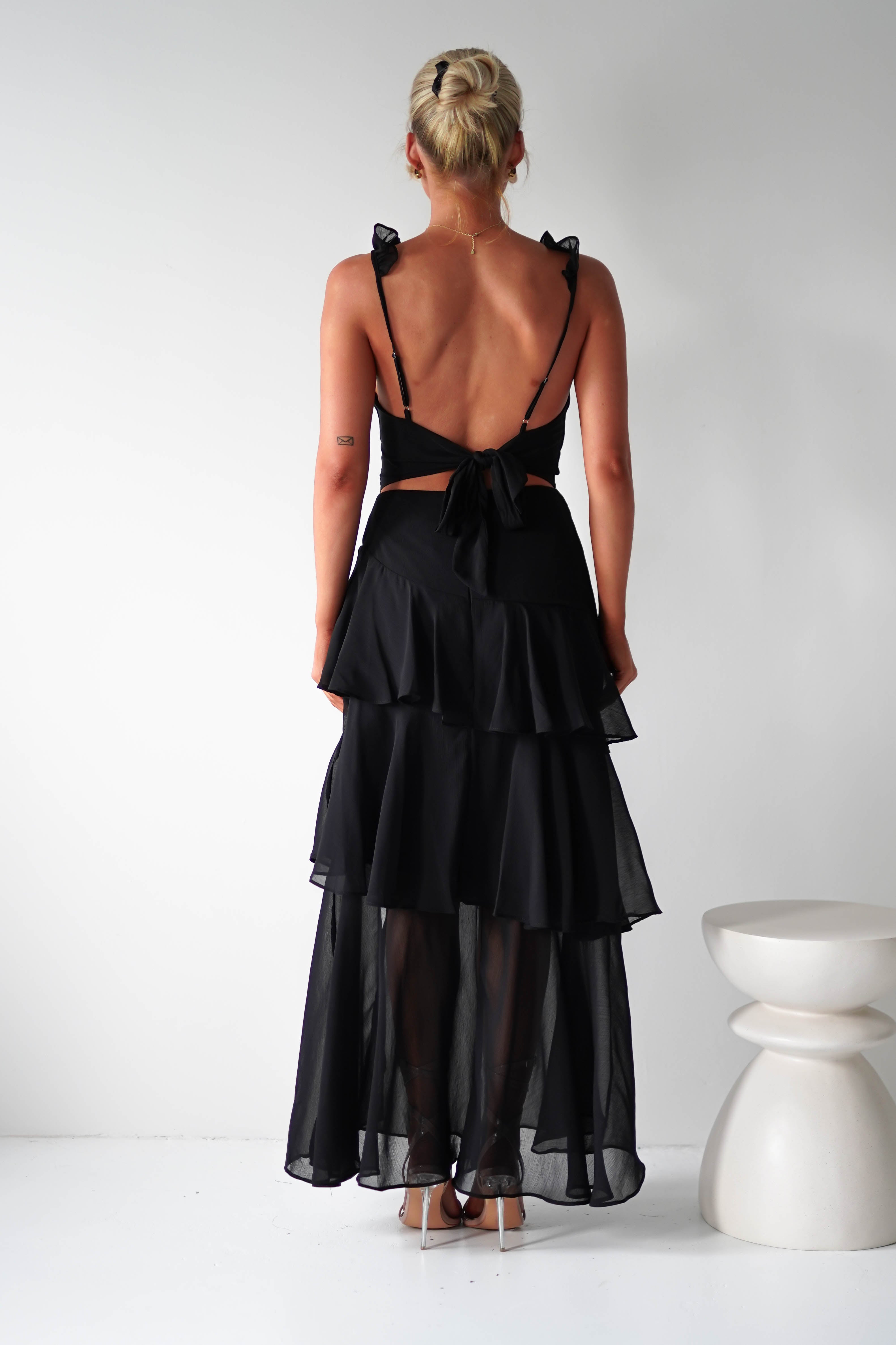 A person with blonde hair in a bun graces a wedding as a guest, donning a black, backless ruffle dress with a tiered sheer skirt of chiffon fabric. They stand facing away, near a white sculptural stool, the dress elegantly tied at the lower back against a plain white backdrop.