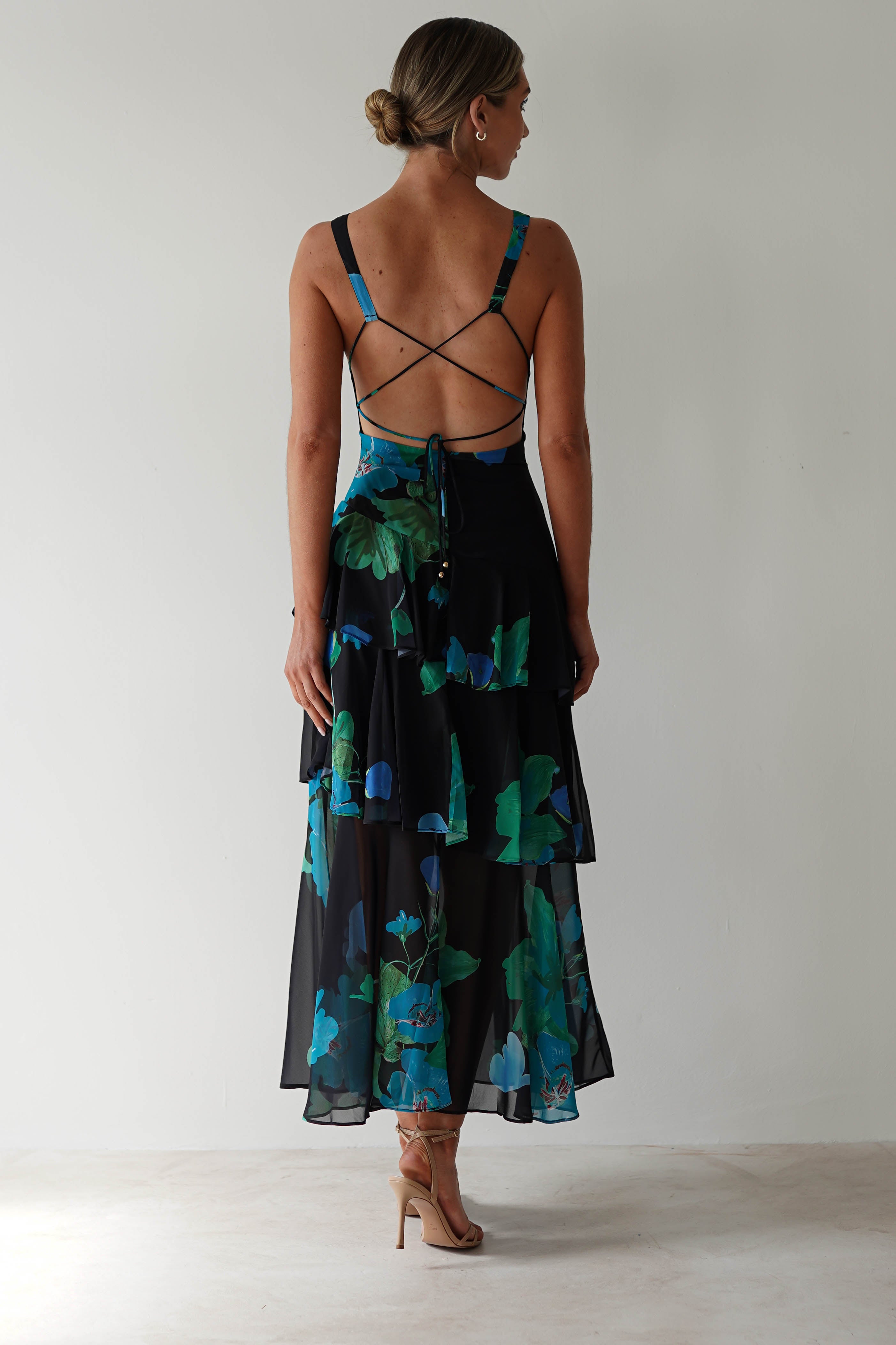 Rear view of a person with hair tied up, wearing a sleeveless black floral midi dress in chiffon fabric, adorned with blue and green floral patterns. The dress features crisscross straps at the back. They stand against a plain background, wearing beige heels—a perfect choice for wedding guest attire.
