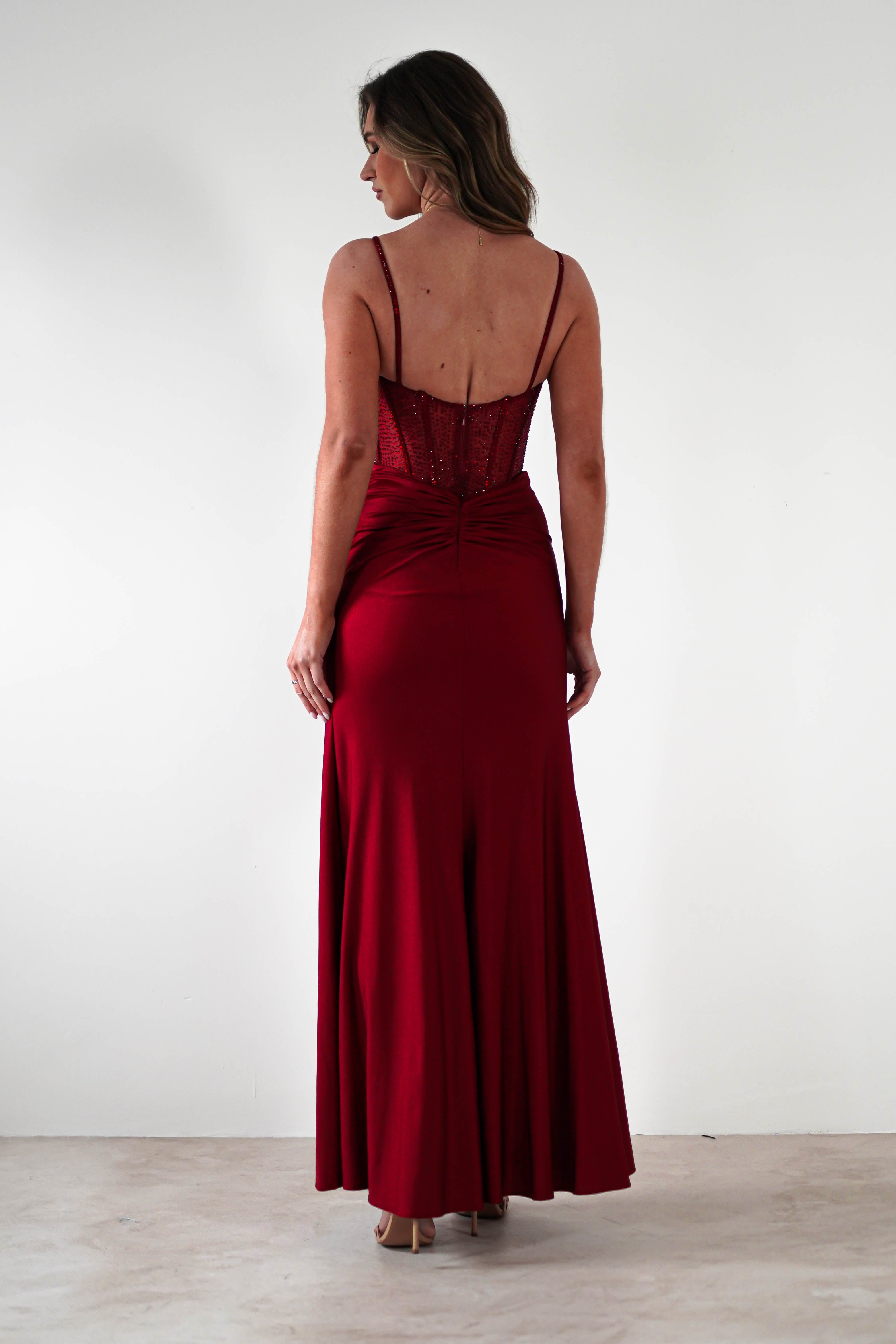 A woman stands facing away from the camera, wearing a floor-length, fitted gown in deep red with thin shoulder straps and a ruched bodice—perfect for formal events. The background is plain white.