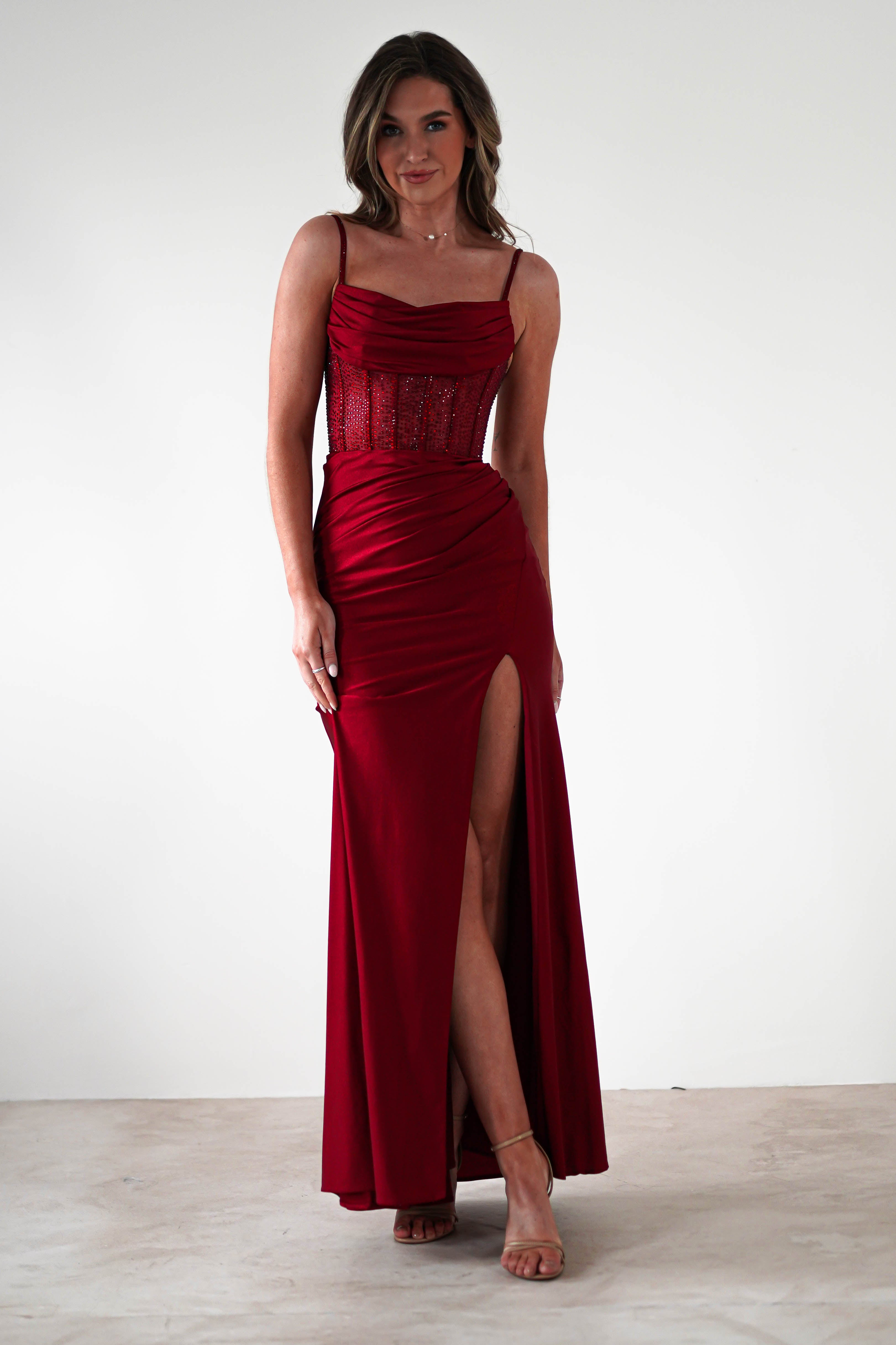 A woman wears a sleeveless, floor-length red fitted gown with a thigh-high slit and rhinestone bodice, standing against a plain white background. She has loose, wavy hair and is wearing strappy heels—perfect for formal events.