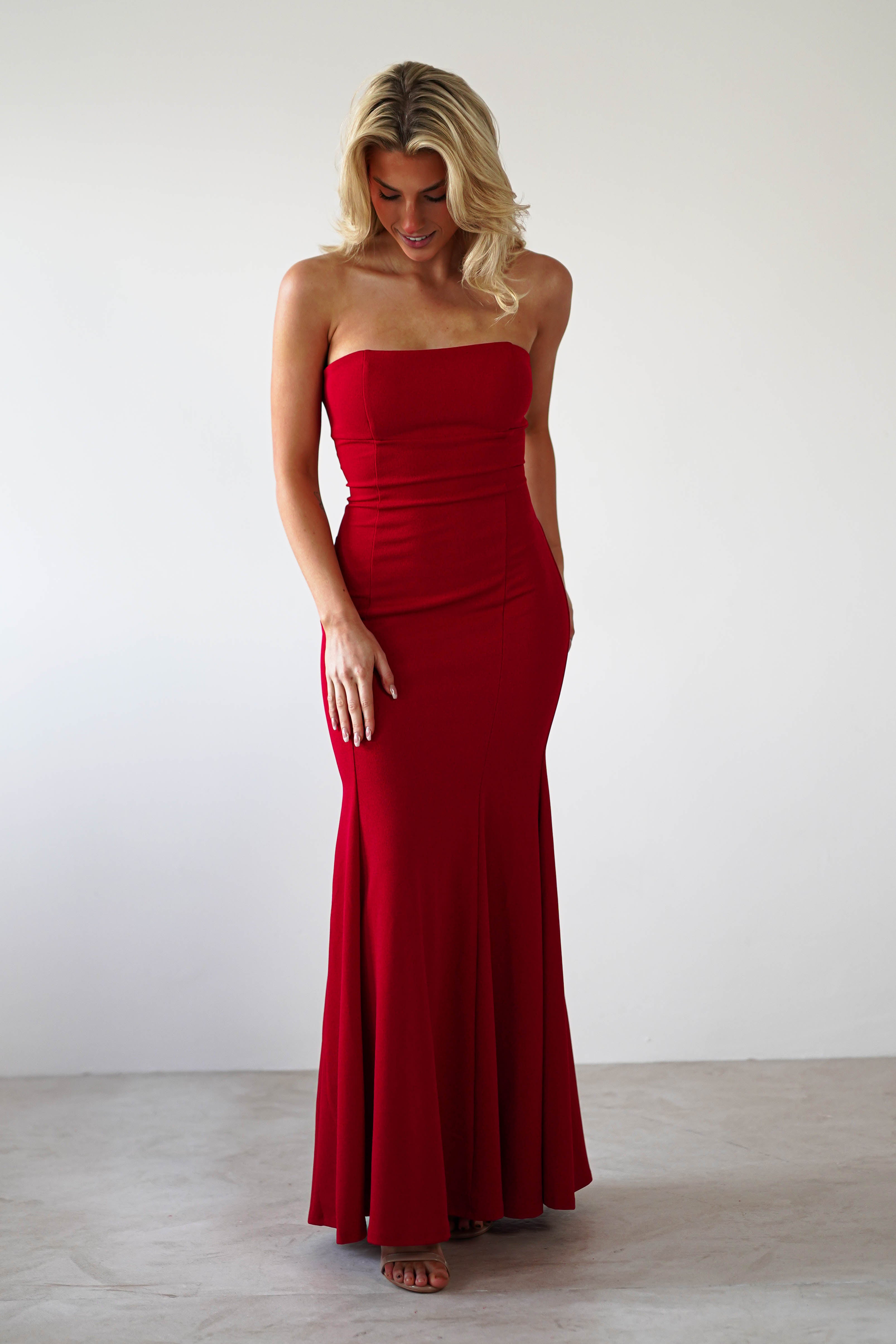 A woman poses in a strapless evening gown with a mermaid silhouette, hands in her dress pockets. The fitted, floor-length red gown complements her long, wavy blonde hair as she gazes downward against a plain white wall.