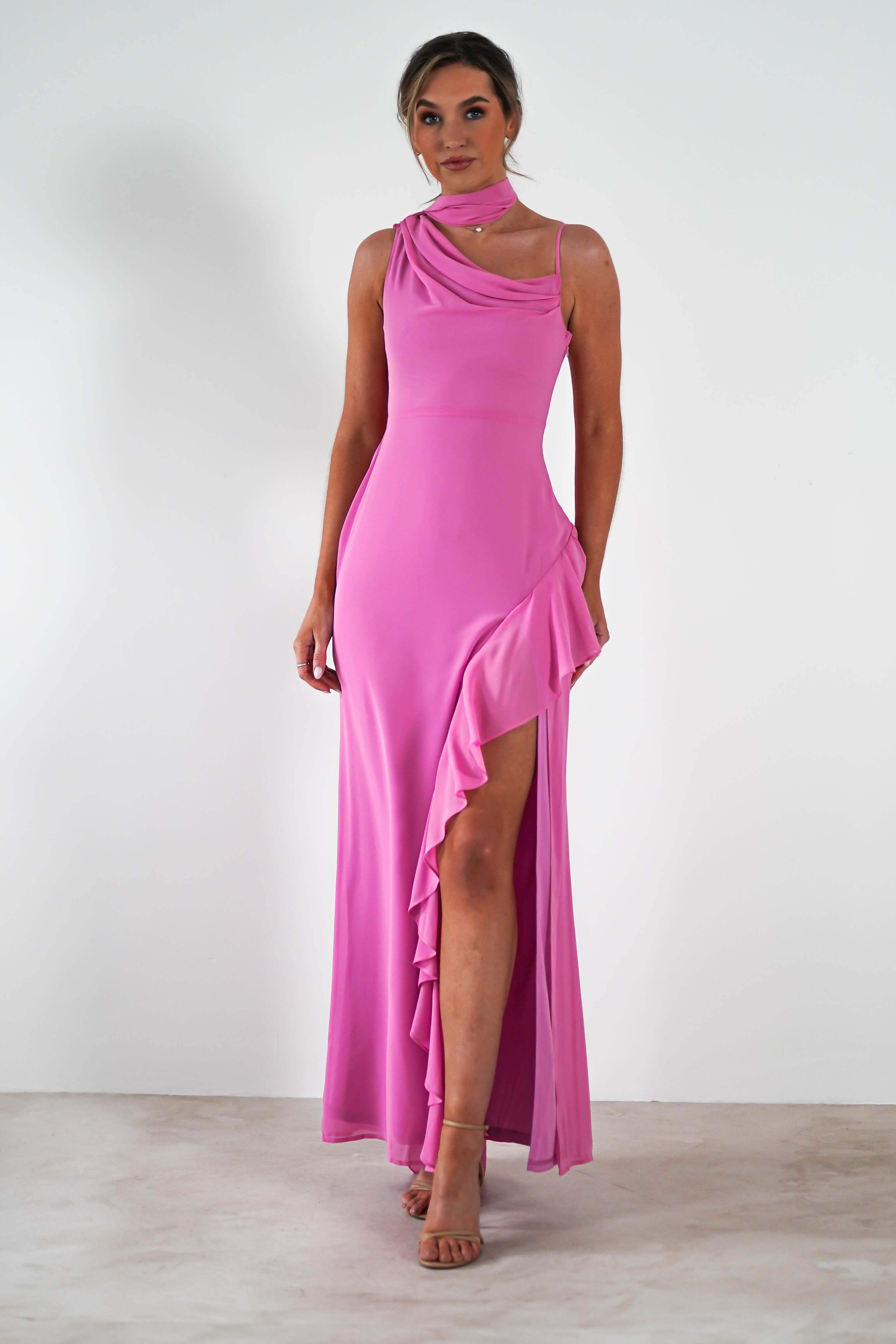 A woman in a flowing coral one-shoulder gown poses against a plain white background. The wedding guest dress features an asymmetrical neckline and a ruffled hem with a thigh-high slit. She stands confidently with one leg slightly forward.