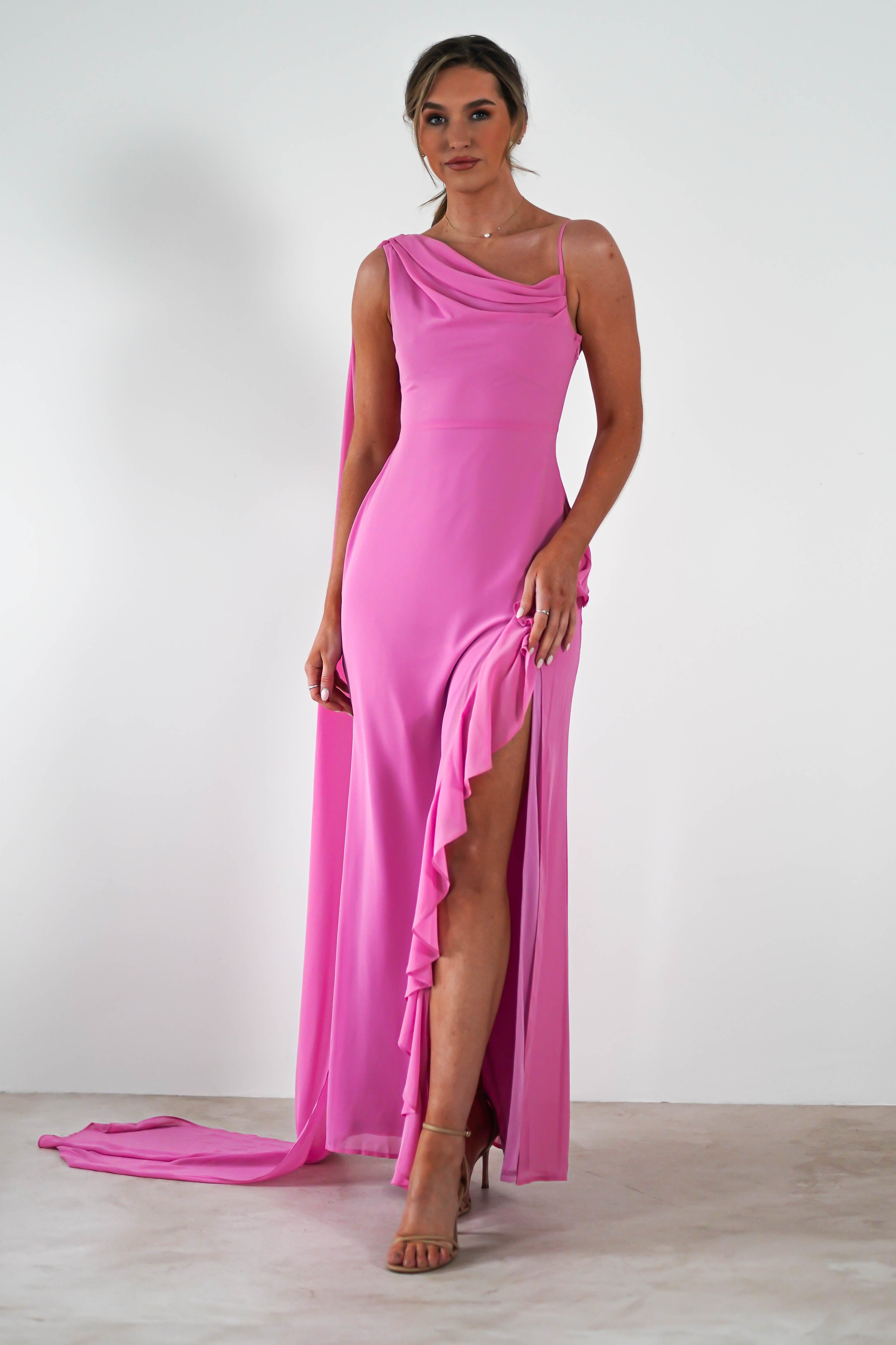 A woman in a coral one-shoulder gown featuring a thigh-high slit and elegant draped neckline poses against a white background. She is wearing high heels, and her hair is styled up with grace.