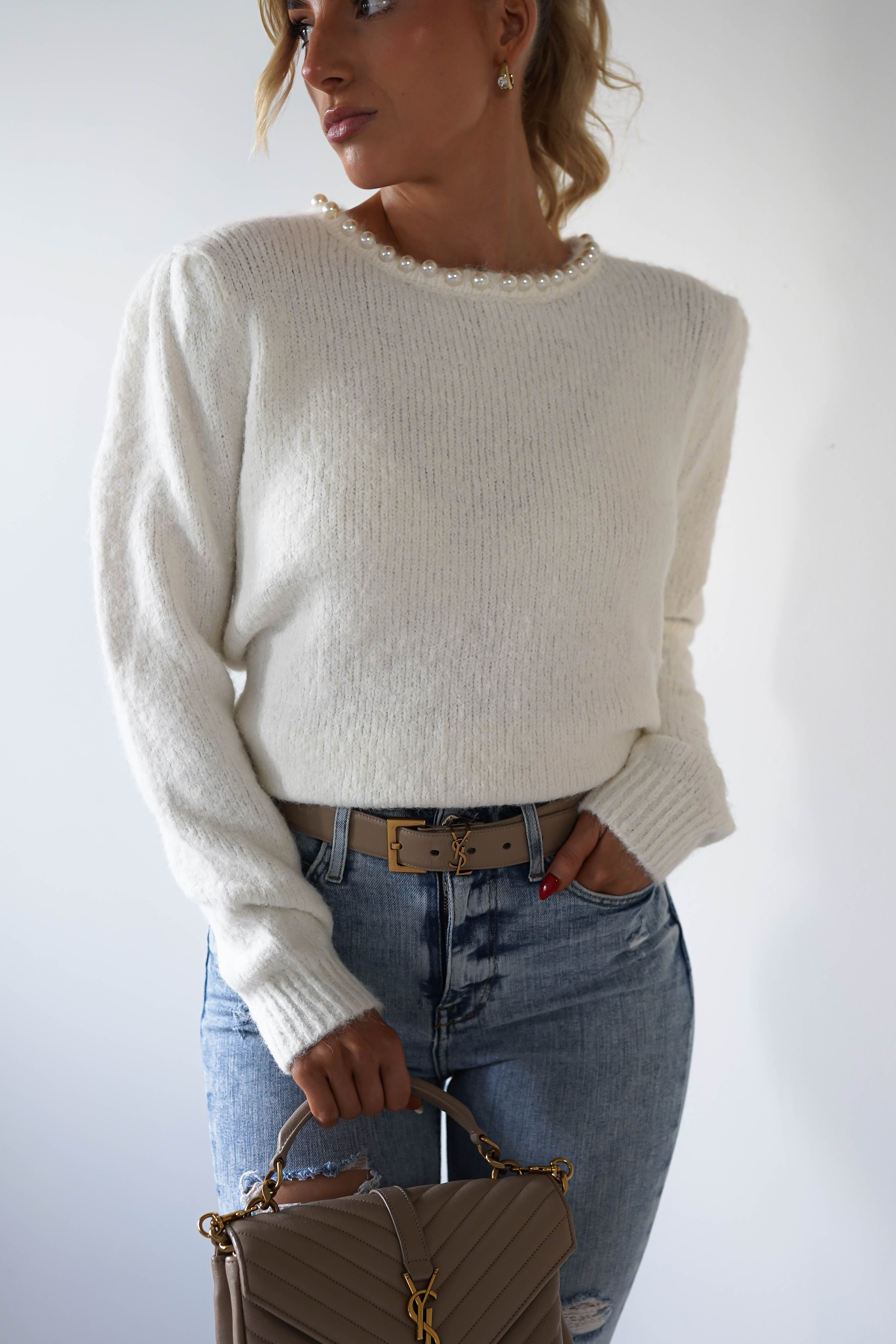 A person wearing a super soft white sweater with pearl embellishments around the neckline and light blue jeans holds a quilted beige handbag. They stand against a plain background, looking to the side with one hand in their pocket.