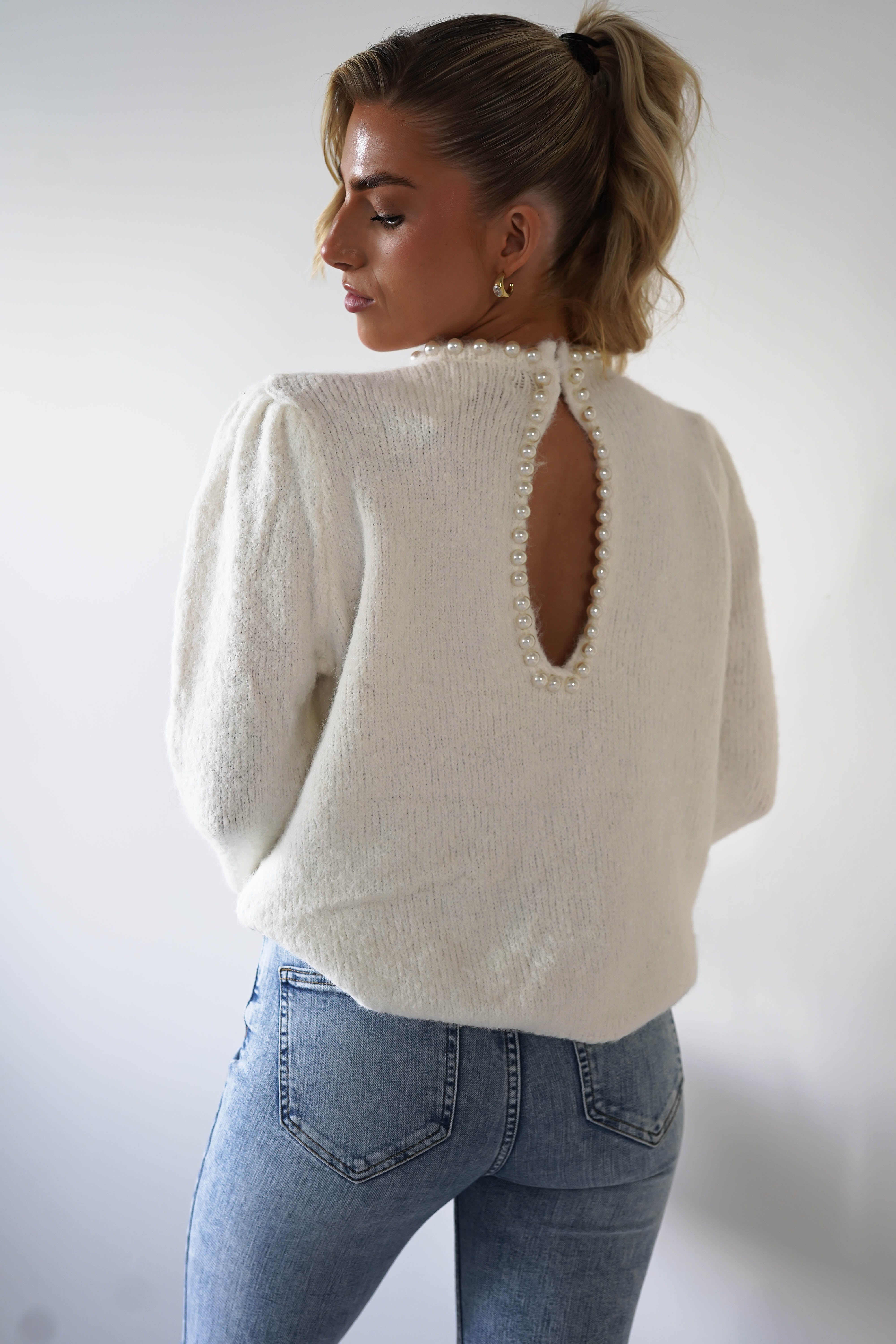 A person with long hair in a ponytail wears a white, super soft jumper with pearl embellishments and an open keyhole detail at the back. Complemented by light blue jeans, they stand against a plain white background, looking over their shoulder.