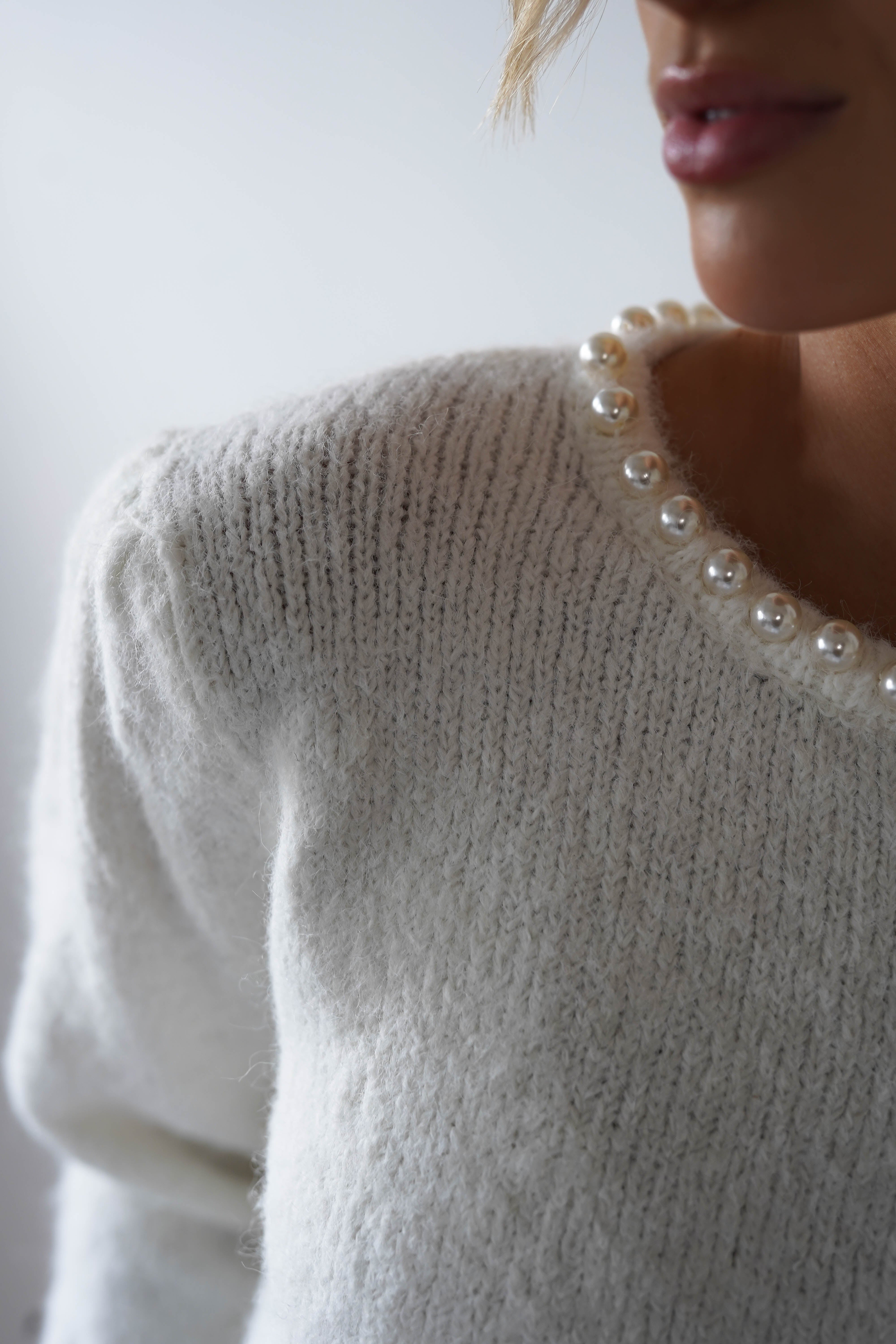 A close-up showcases a person in a cream-colored super soft jumper adorned with large, decorative pearl embellishments around the neckline. The background is softly blurred, revealing only part of the persons face.