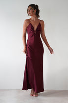 Woman wearing Adley Soft Satin Maxi Dress in burgundy with plunging neckline, thin spaghetti straps, and floor-length satin finish, styled with nude heels.