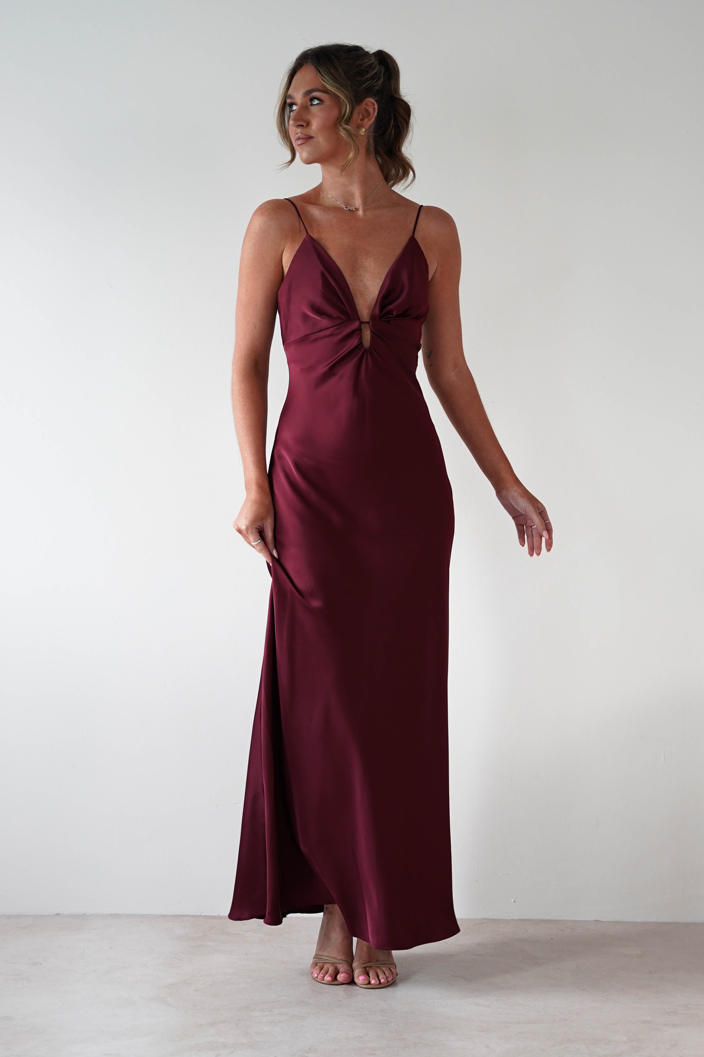 Woman wearing Adley Soft Satin Maxi Dress in burgundy with plunging neckline, thin spaghetti straps, and floor-length satin finish, styled with nude heels.