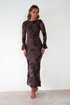 Woman wearing a brown floral embroidered Amelita maxi dress with long sleeves and fitted silhouette, styled with nude heels against a white background.
