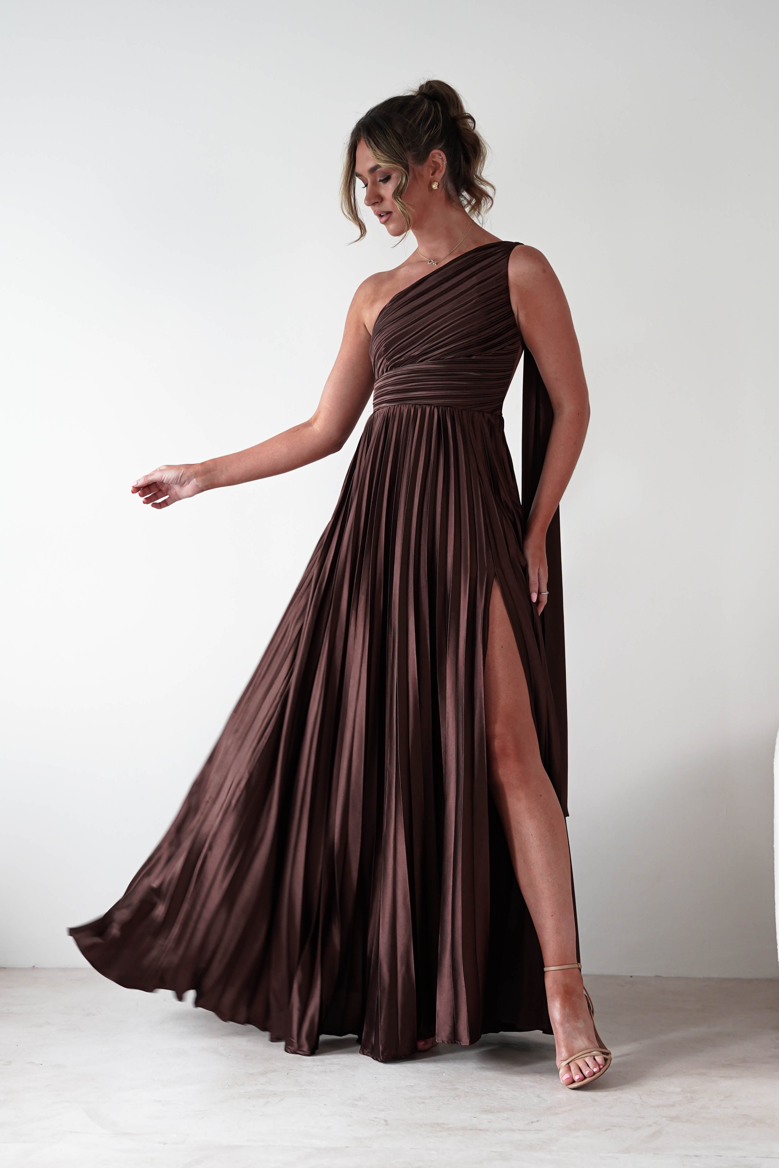 Woman wearing Jianna Pleated Maxi Dress in brown with one-shoulder neckline, flowing pleated skirt, and thigh-high slit, styled with nude strappy heels.