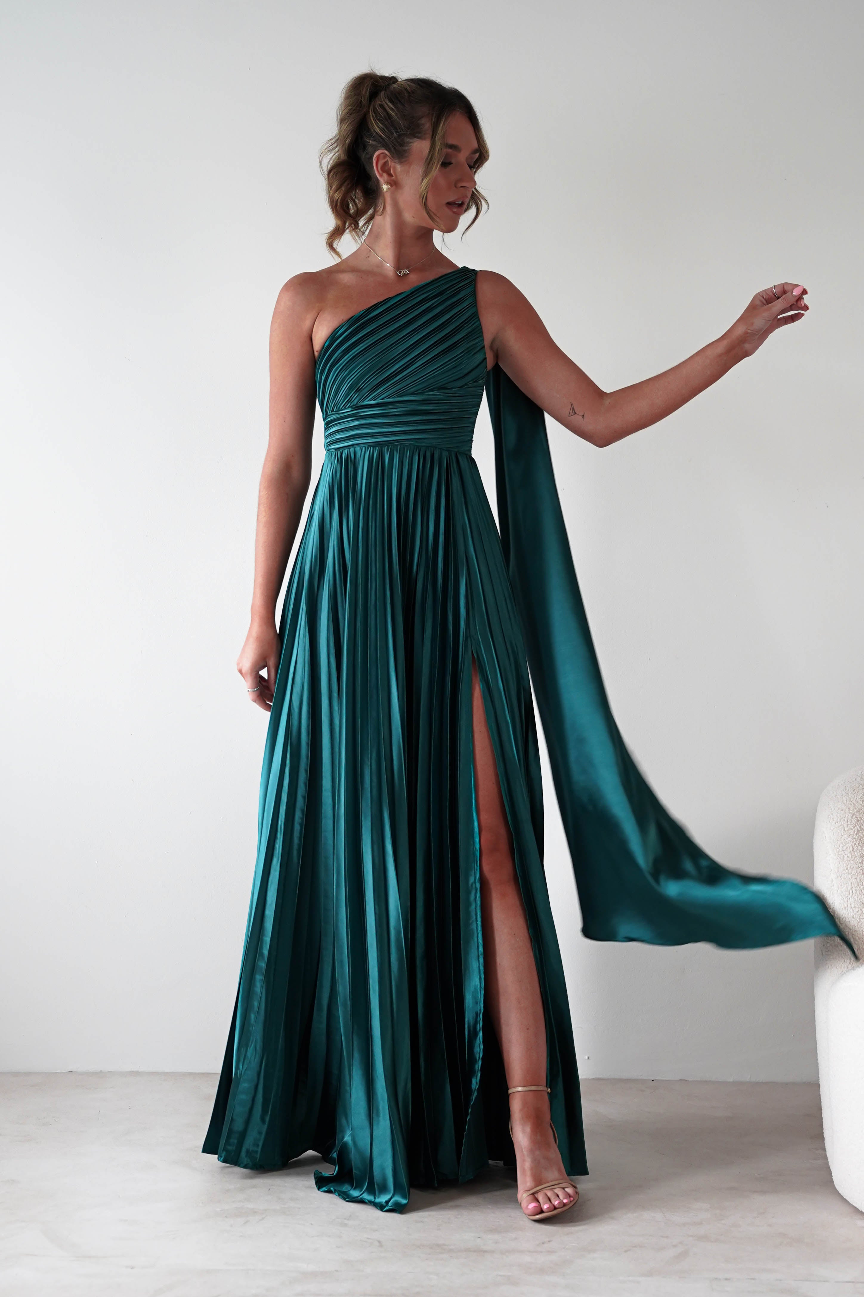 Woman wearing Jianna Pleated Maxi Dress in emerald green with one-shoulder neckline, draped sash detail, pleated skirt, and thigh-high slit, styled with nude heels.
