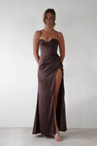 Woman wearing Julianne Corset Satin Gown in brown with draped neckline, corset-style bodice, and high leg slit, styled with nude heels against a minimal backdrop.