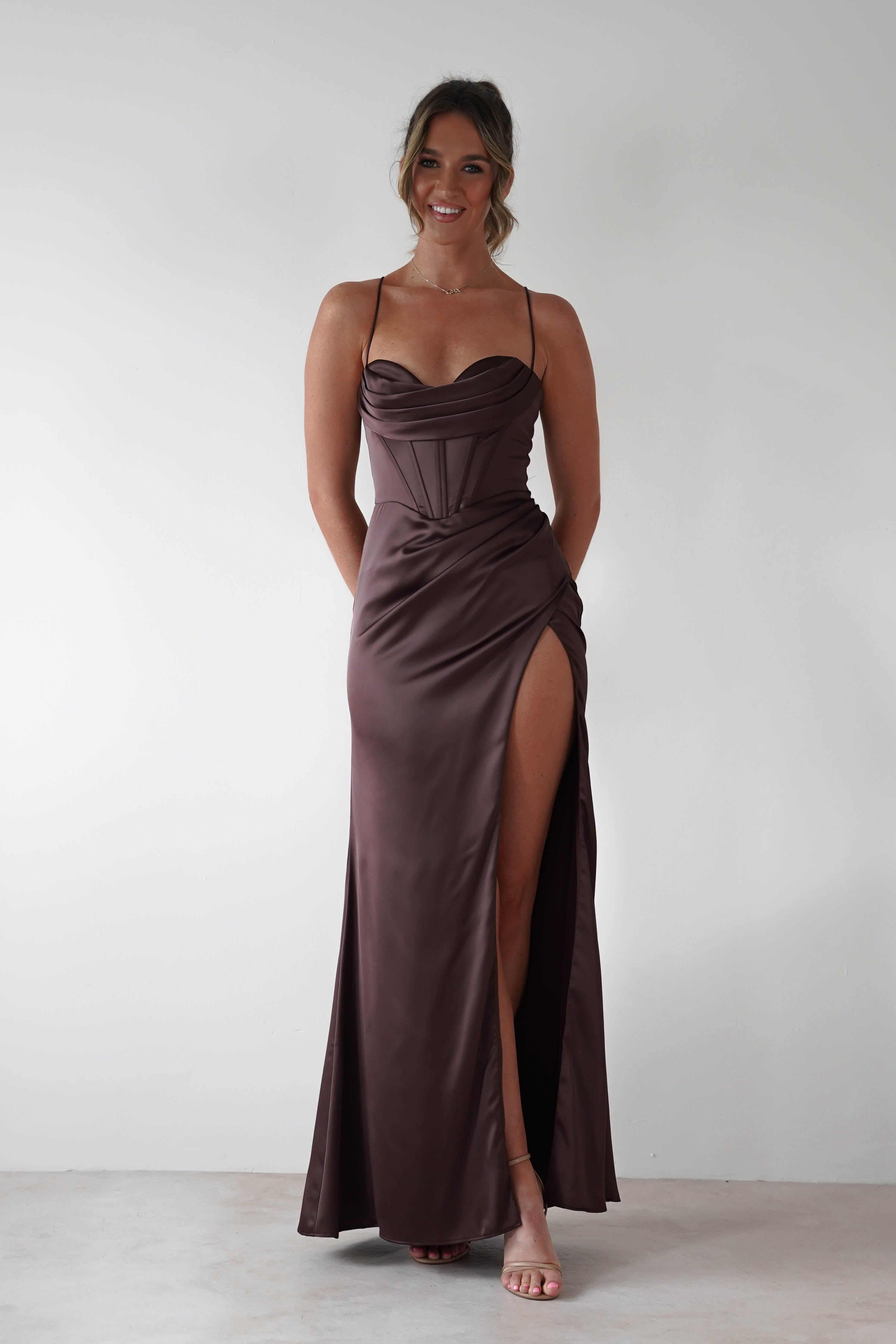 Woman wearing Julianne Corset Satin Gown in brown with draped neckline, corset-style bodice, and high leg slit, styled with nude heels against a minimal backdrop.