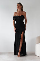 Woman wearing the Karlene black off-the-shoulder gown with a satin fold-over neckline and thigh-high slit, styled with gold earrings and nude heels.