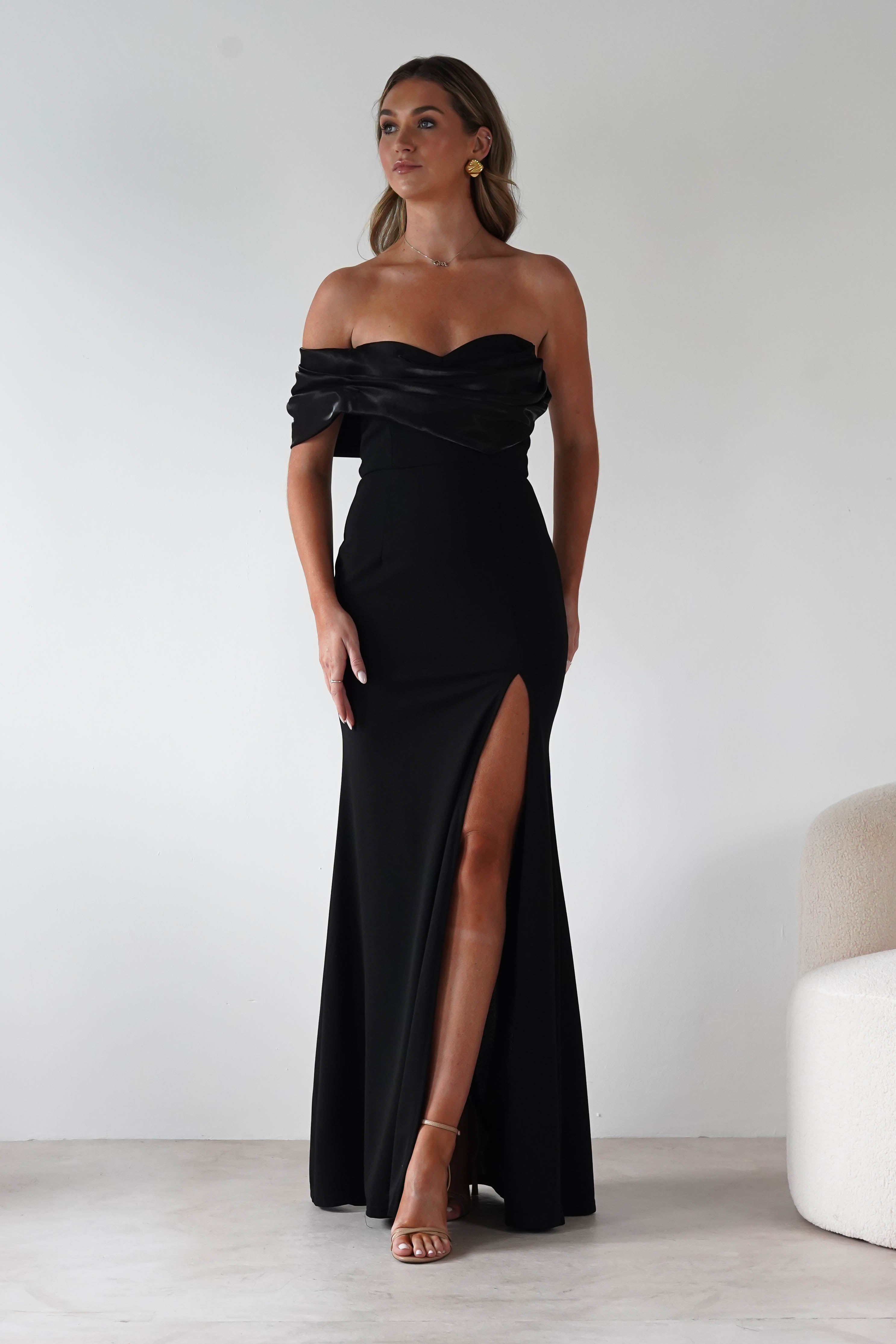 Woman wearing the Karlene black off-the-shoulder gown with a satin fold-over neckline and thigh-high slit, styled with gold earrings and nude heels.