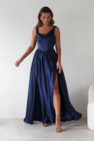 Woman wearing the Katalia navy satin gown with a draped neckline and thigh-high slit, paired with gold earrings and nude heels.