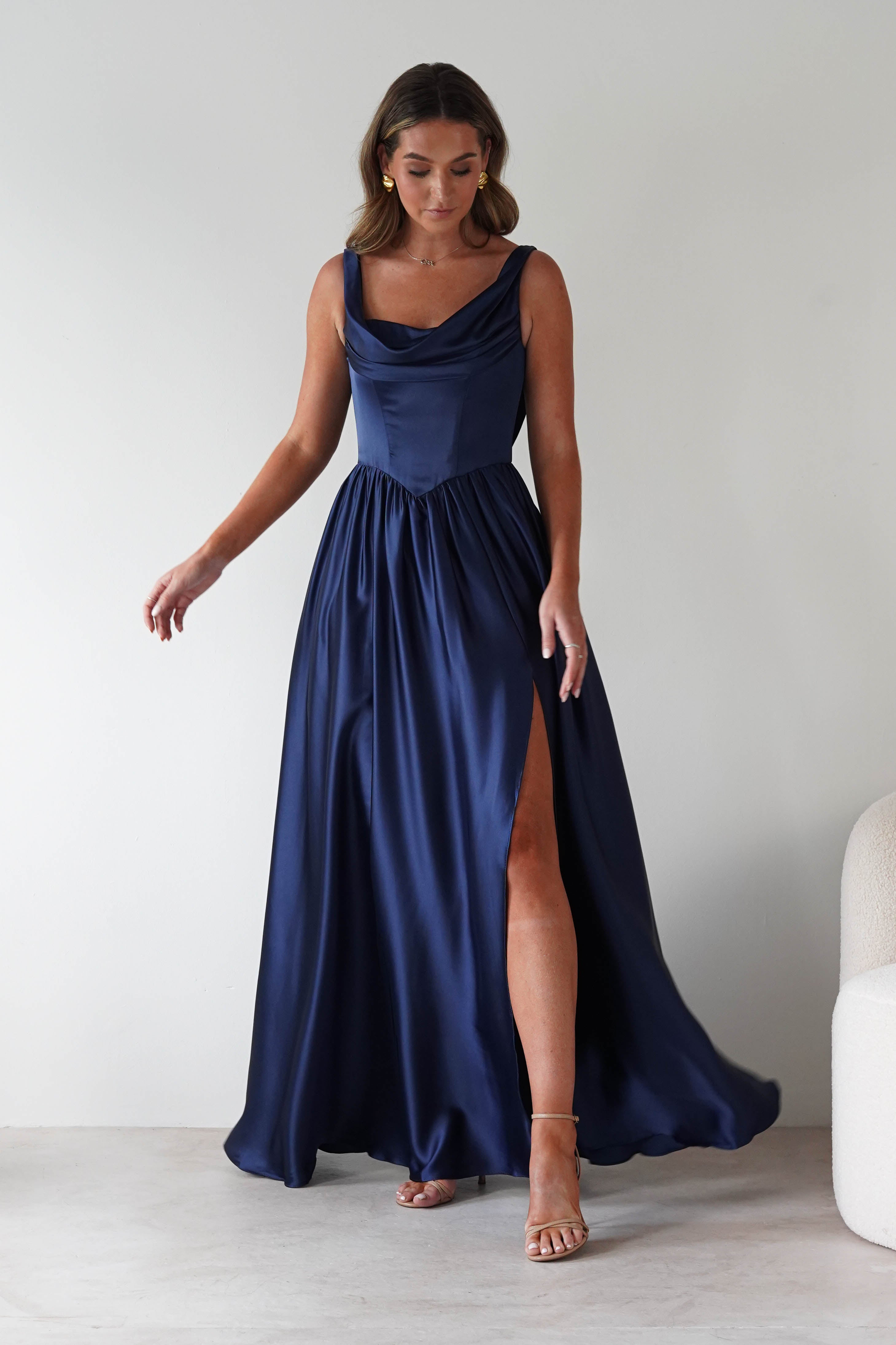 Woman wearing the Katalia navy satin gown with a draped neckline and thigh-high slit, paired with gold earrings and nude heels.