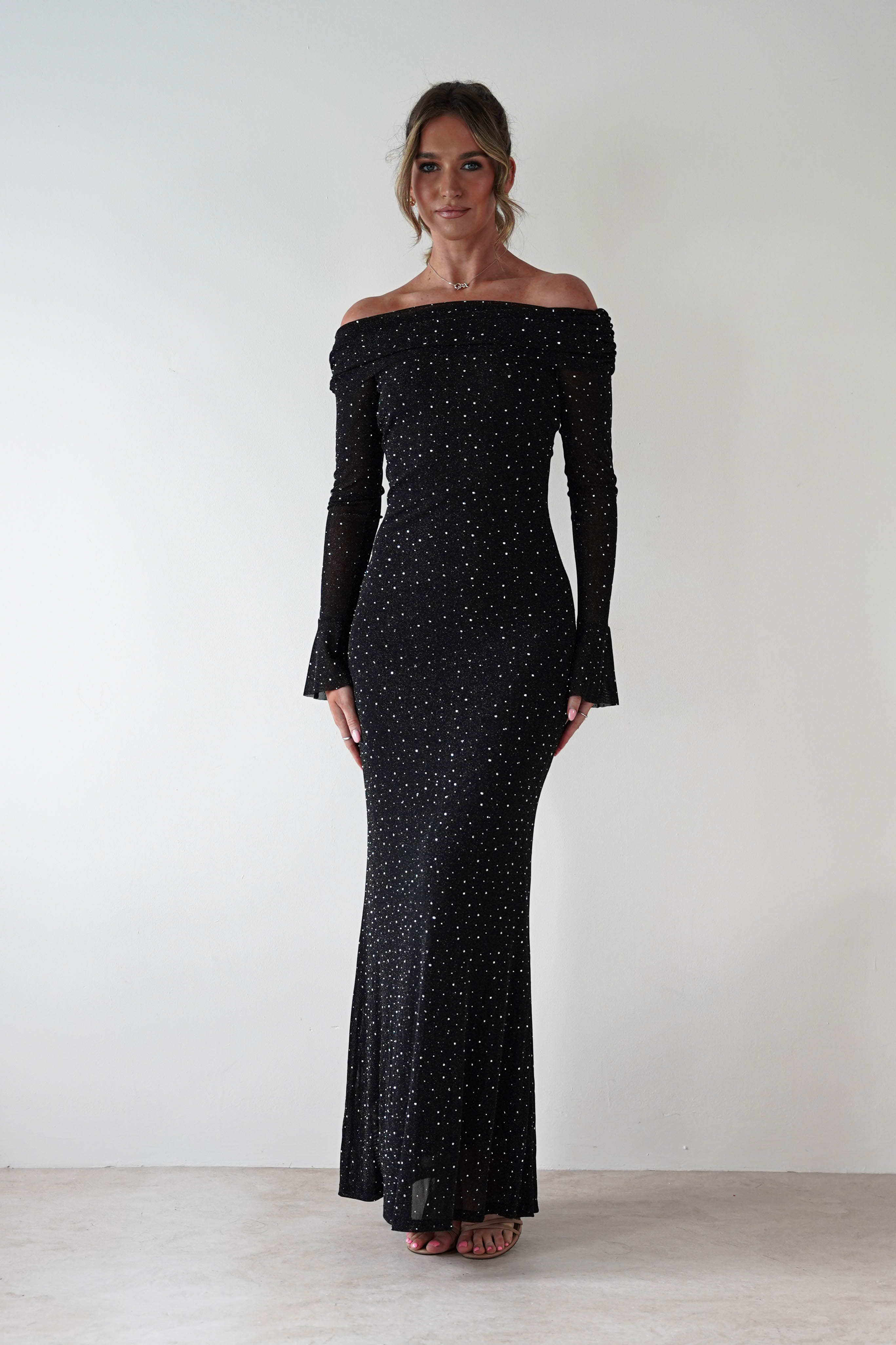 Woman wearing Katalina Sparkling Rhinestone Maxi Dress in black with off-shoulder neckline, long sleeves, and rhinestone embellishments throughout the fitted silhouette.