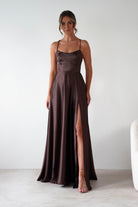 Woman wearing Leelie Silky Satin Maxi Gown in brown with a fitted bodice, thin spaghetti straps, flowing skirt, and high slit, paired with nude heels.