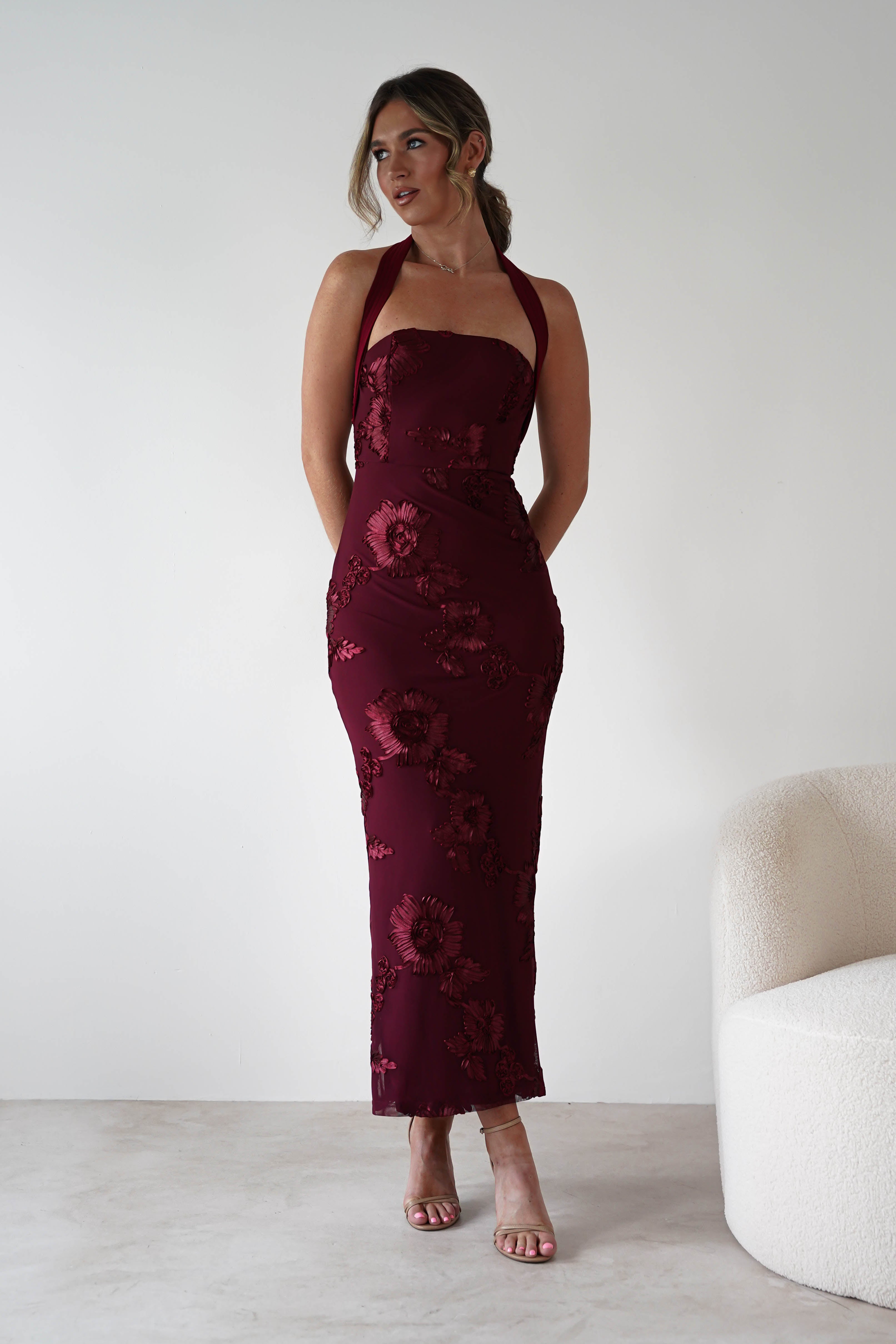 Woman wearing Lilani Textured Midaxi Dress in wine with halter neckline, embroidered floral detailing, and a body-hugging midaxi length, styled with nude heels.