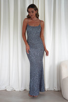 Woman wearing a silver sequin Maria bodycon maxi gown with thin straps and fitted silhouette, styled with nude heels in front of white curtains.