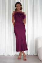 Woman wearing a plum silky satin Neve slip dress with feather detailing on the neckline, fitted silhouette, and nude heels against white curtains.