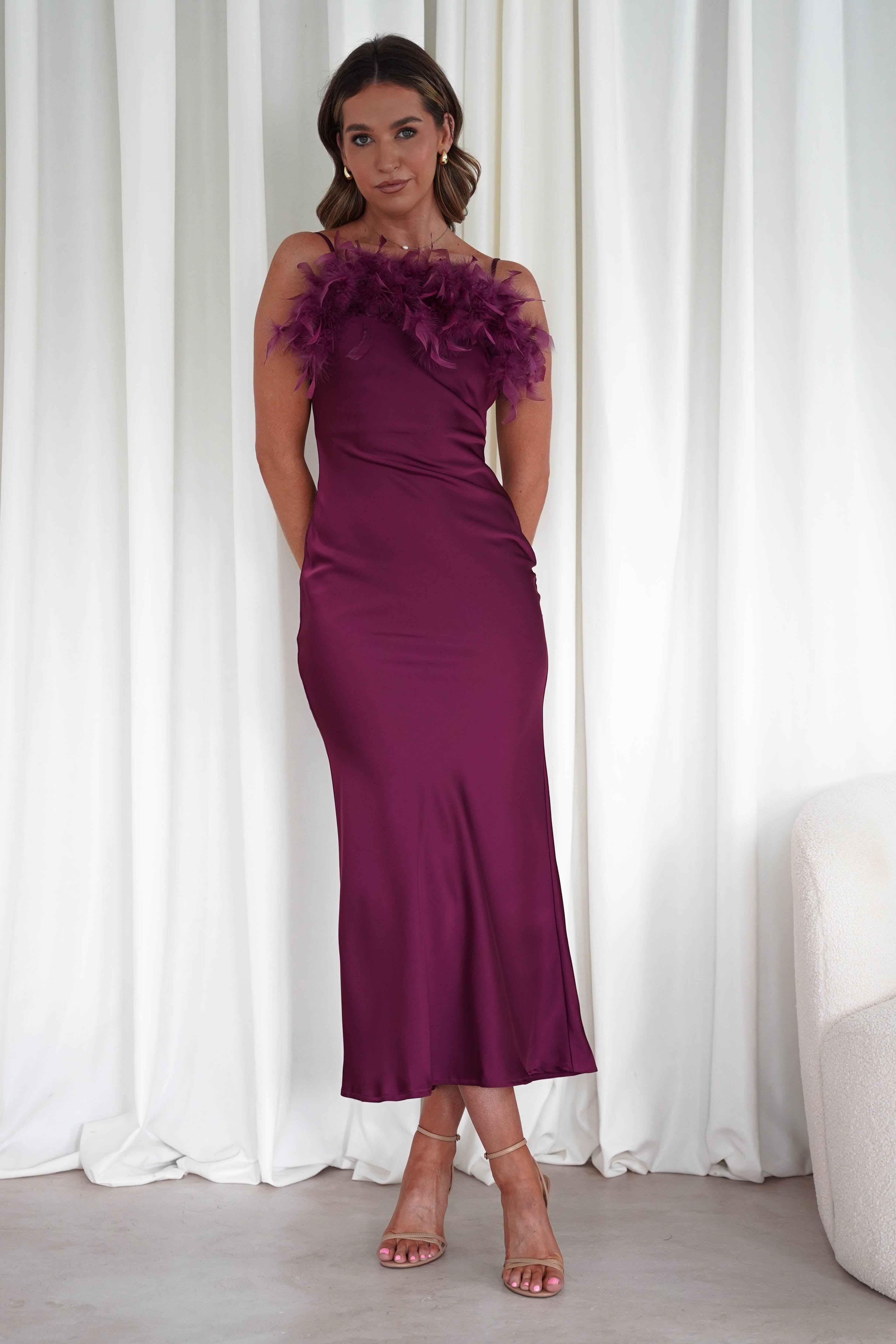 Woman wearing a plum silky satin Neve slip dress with feather detailing on the neckline, fitted silhouette, and nude heels against white curtains.