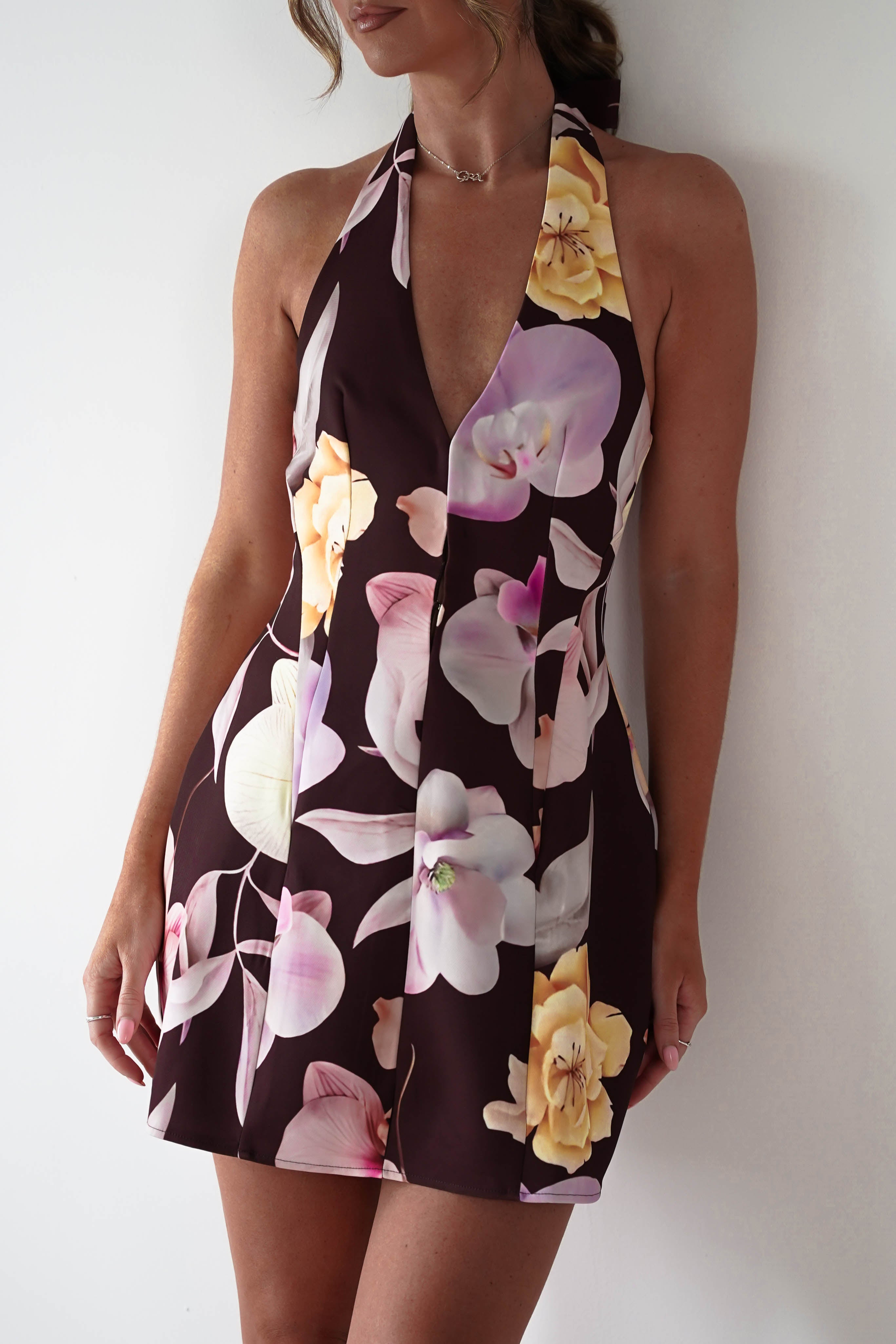 Woman wearing Peony Structured Halter Neck Mini Dress in brown with a plunging neckline, bold pink and yellow floral print, and a fitted, tailored silhouette.