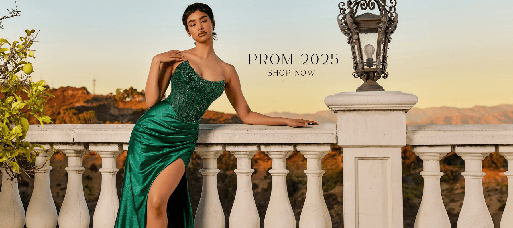 Elegant woman in a green sequined prom dress posing on a white stone terrace at sunset with mountains in the background, featuring the text ‘Prom 2025 – Shop Now