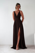 Woman wearing Rebecca Chiffon Maxi Dress in brown with deep V-neckline, fitted waist, flowing chiffon skirt, and thigh-high slit, styled with nude heels.