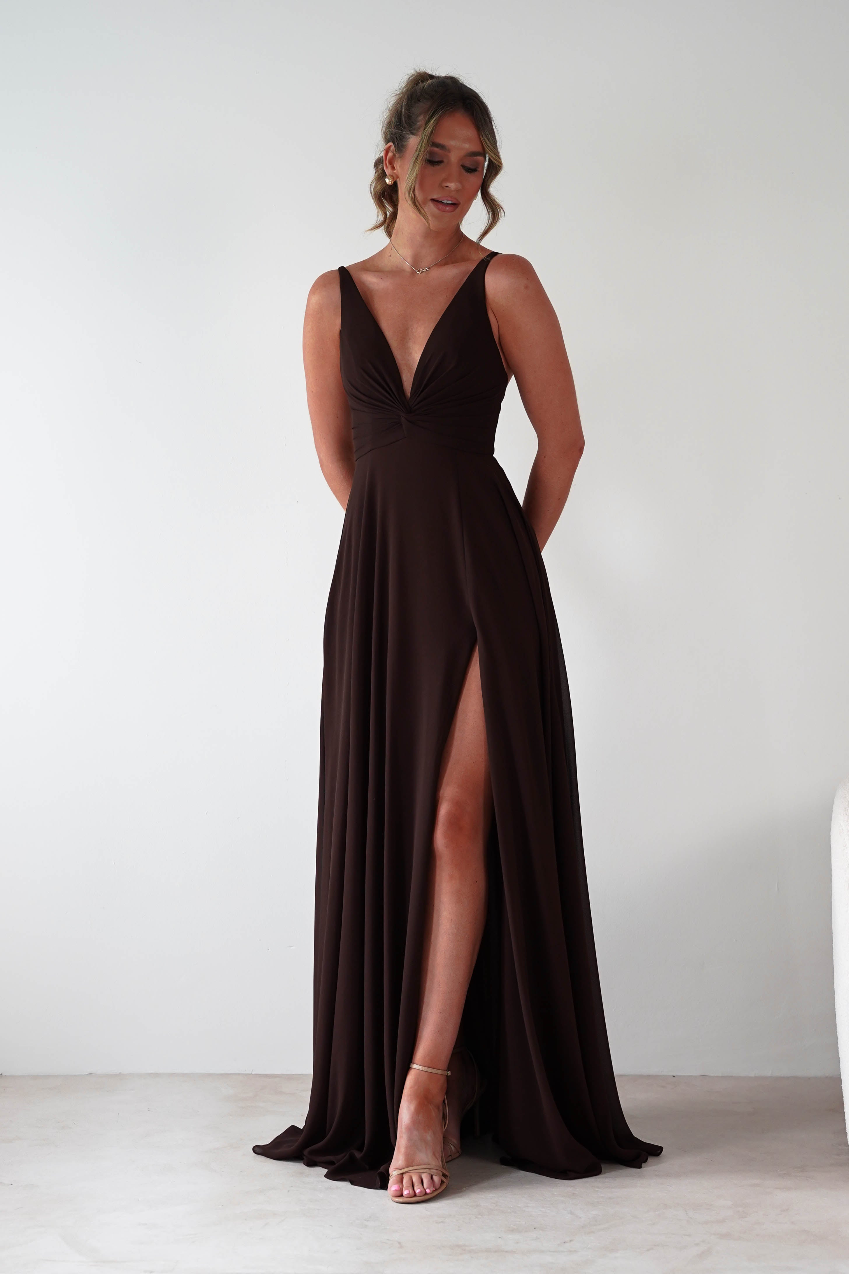 Woman wearing Rebecca Chiffon Maxi Dress in brown with deep V-neckline, fitted waist, flowing chiffon skirt, and thigh-high slit, styled with nude heels.