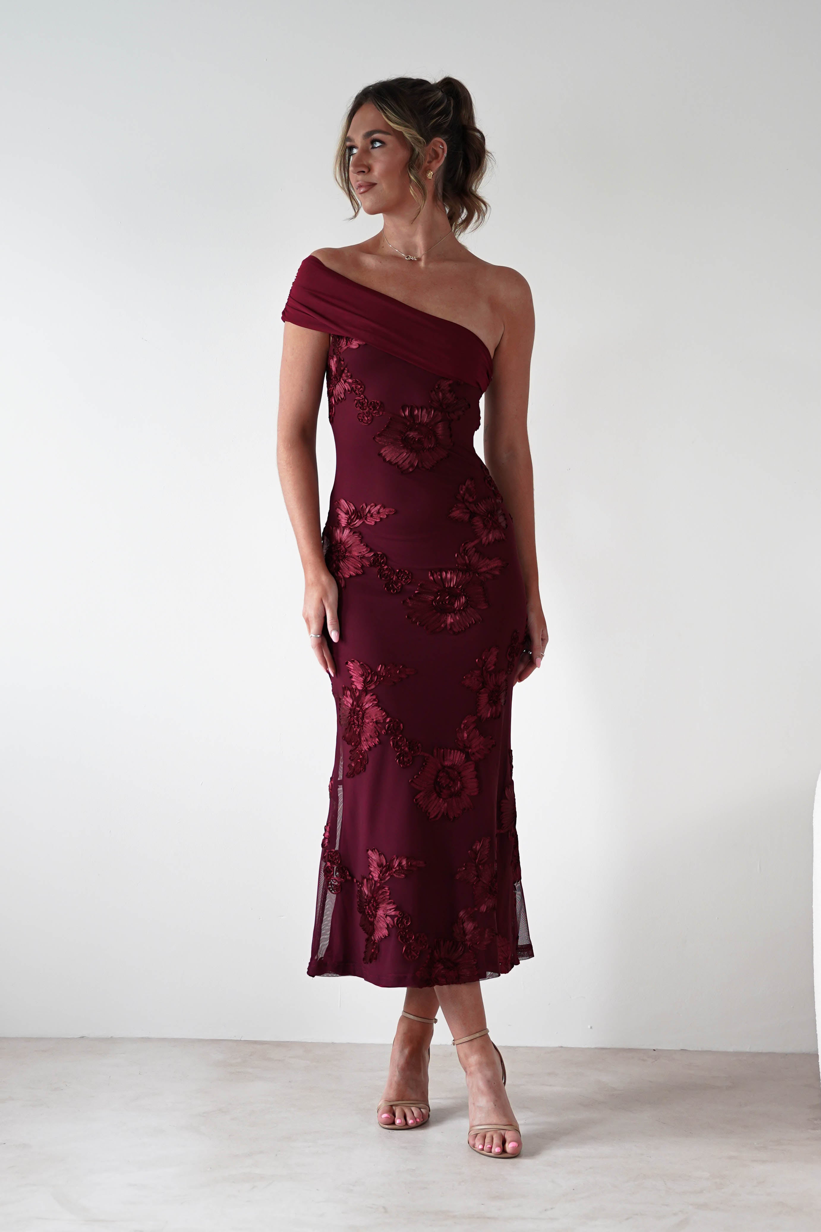 Woman wearing Rochelle Off The Shoulder Midi Dress in wine with floral embroidery, fitted silhouette, and asymmetrical draped neckline, styled with nude heels.