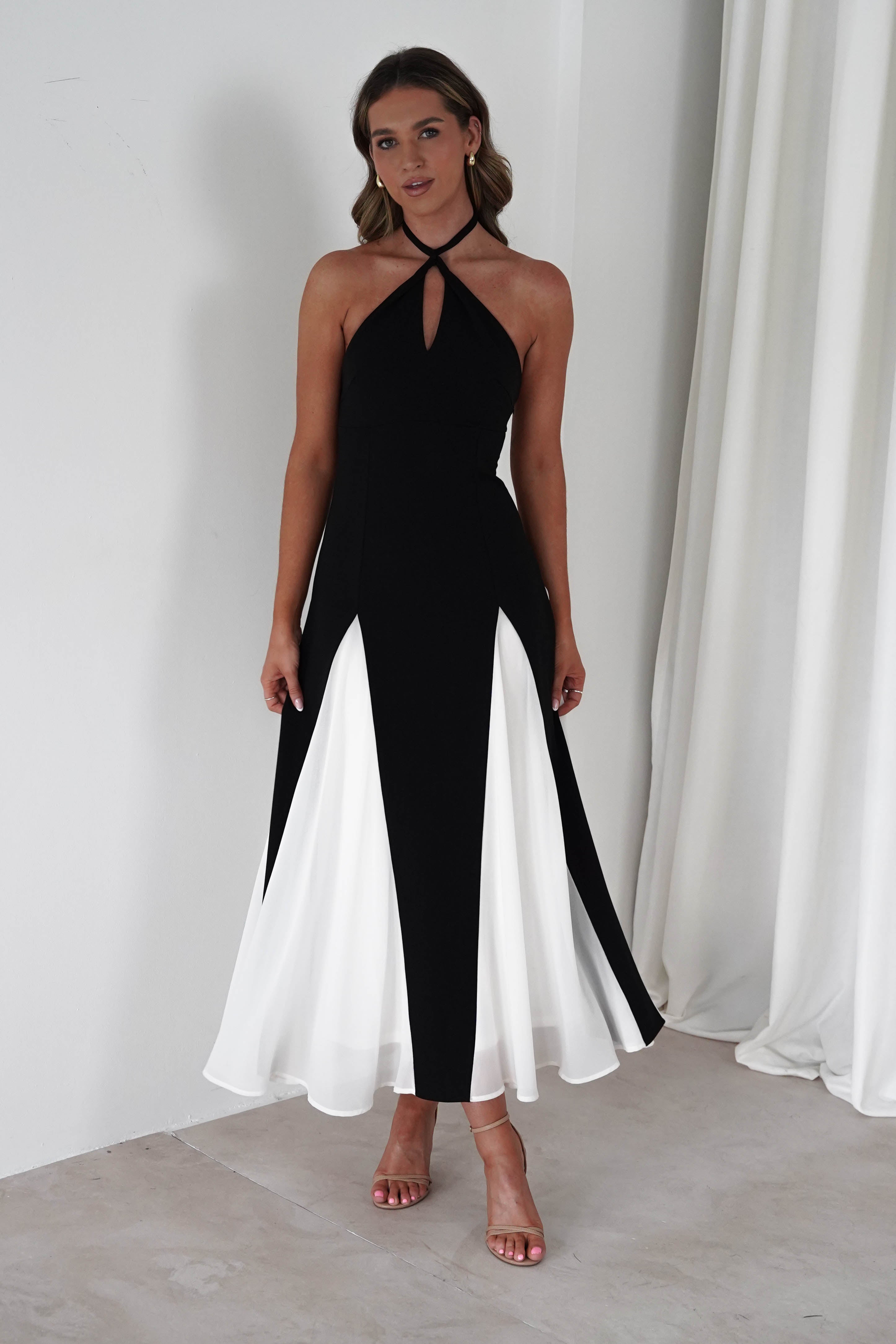 Woman wearing a black and white halter neck Silvana midi dress with keyhole front and flowing panels, paired with nude heels in a minimalist setting.