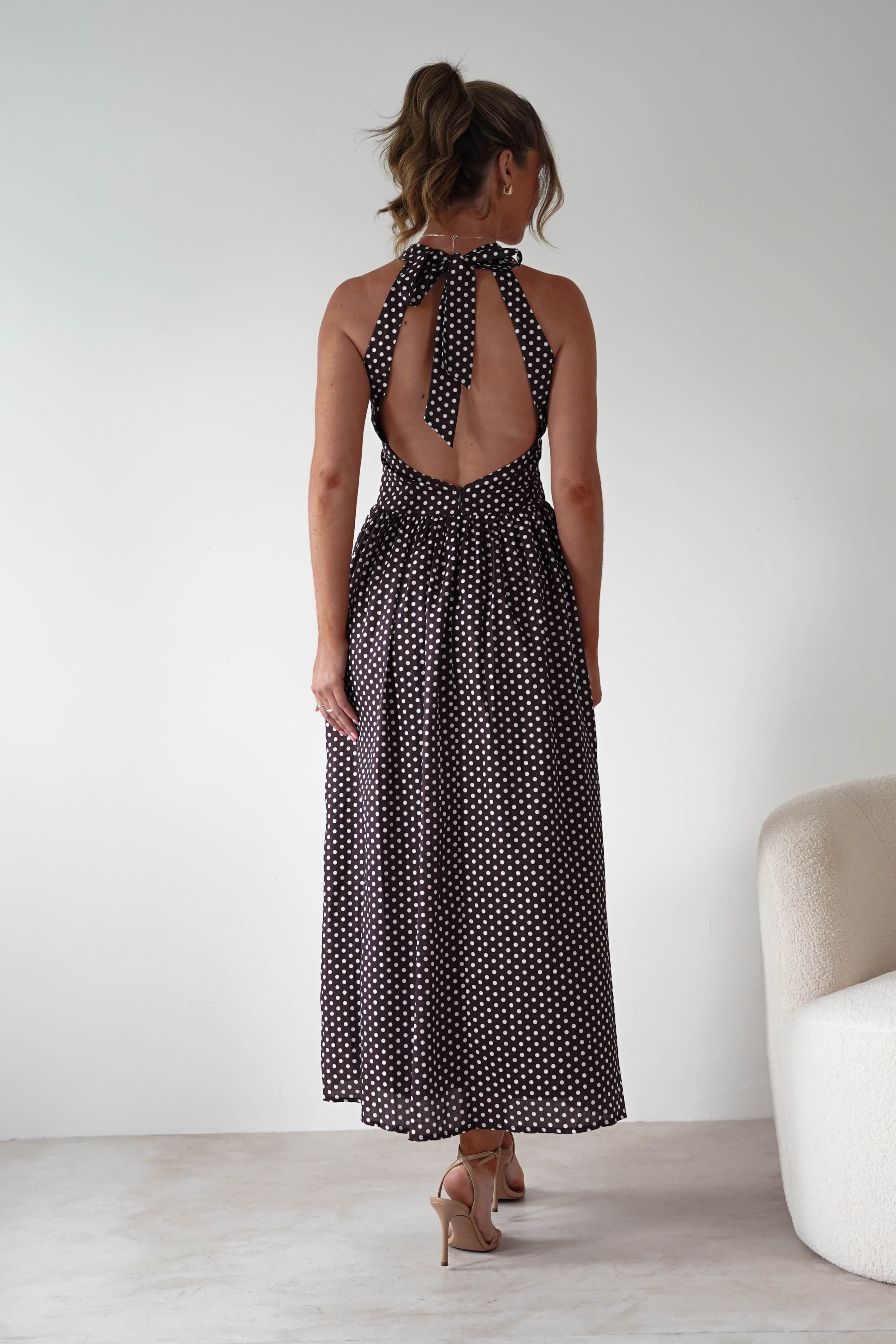 Woman wearing Sivana Polka Dot Skater Maxi Dress in brown with white polka dots, halter tie-back neckline, open back, and flowy maxi skirt styled with nude heels.