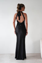 Woman wearing Taylee Halterneck Maxi Dress in black with satin finish, halter tie-back neckline, open back, and floor-length fitted silhouette.