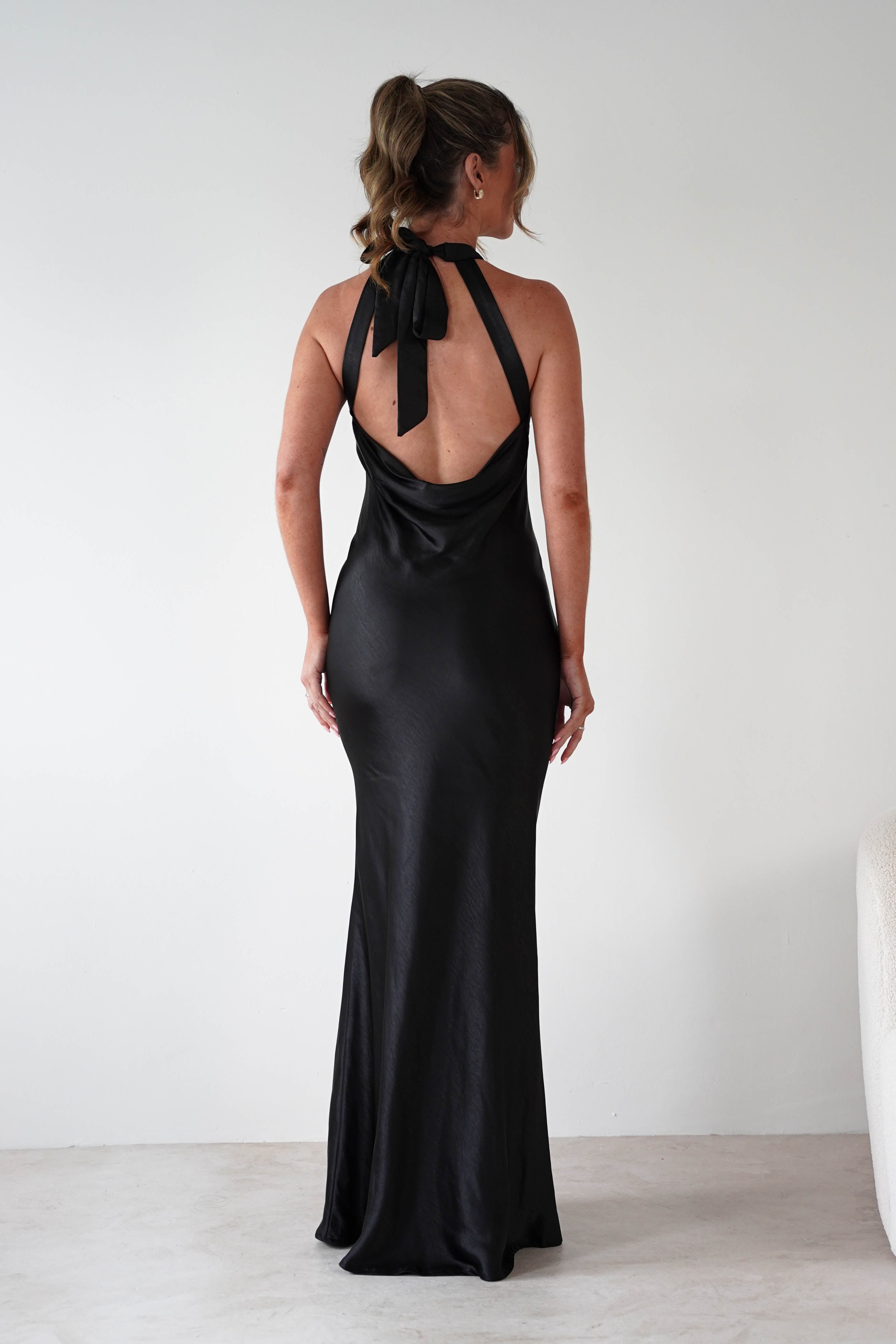 Woman wearing Taylee Halterneck Maxi Dress in black with satin finish, halter tie-back neckline, open back, and floor-length fitted silhouette.