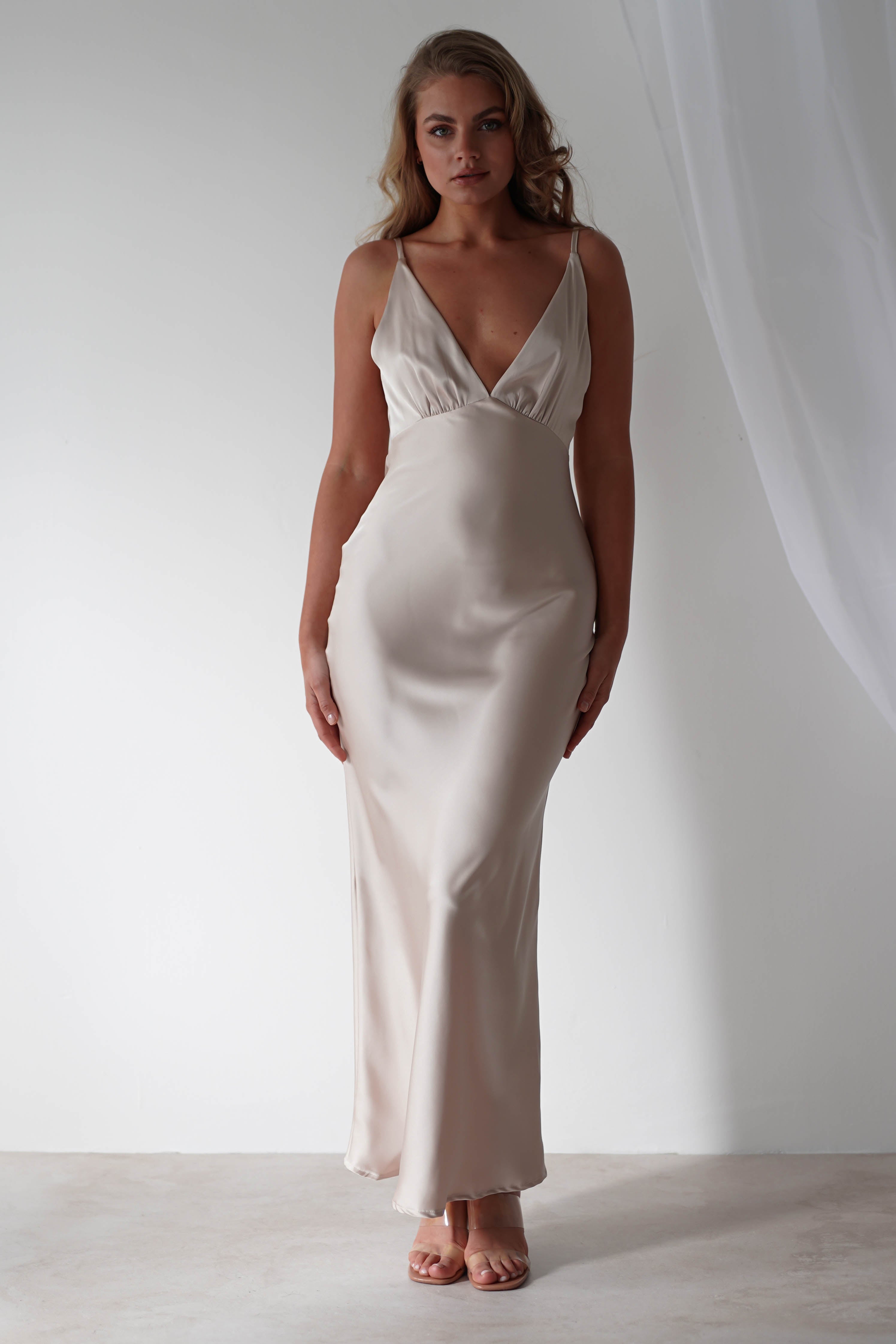 A person wearing a sleek, floor-length satin maxi dress stands against a neutral background. The gown features a deep v-neckline and thin straps. With long, wavy hair, they gaze directly at the camera, ready to dazzle as a bridesmaid or elegant wedding guest.