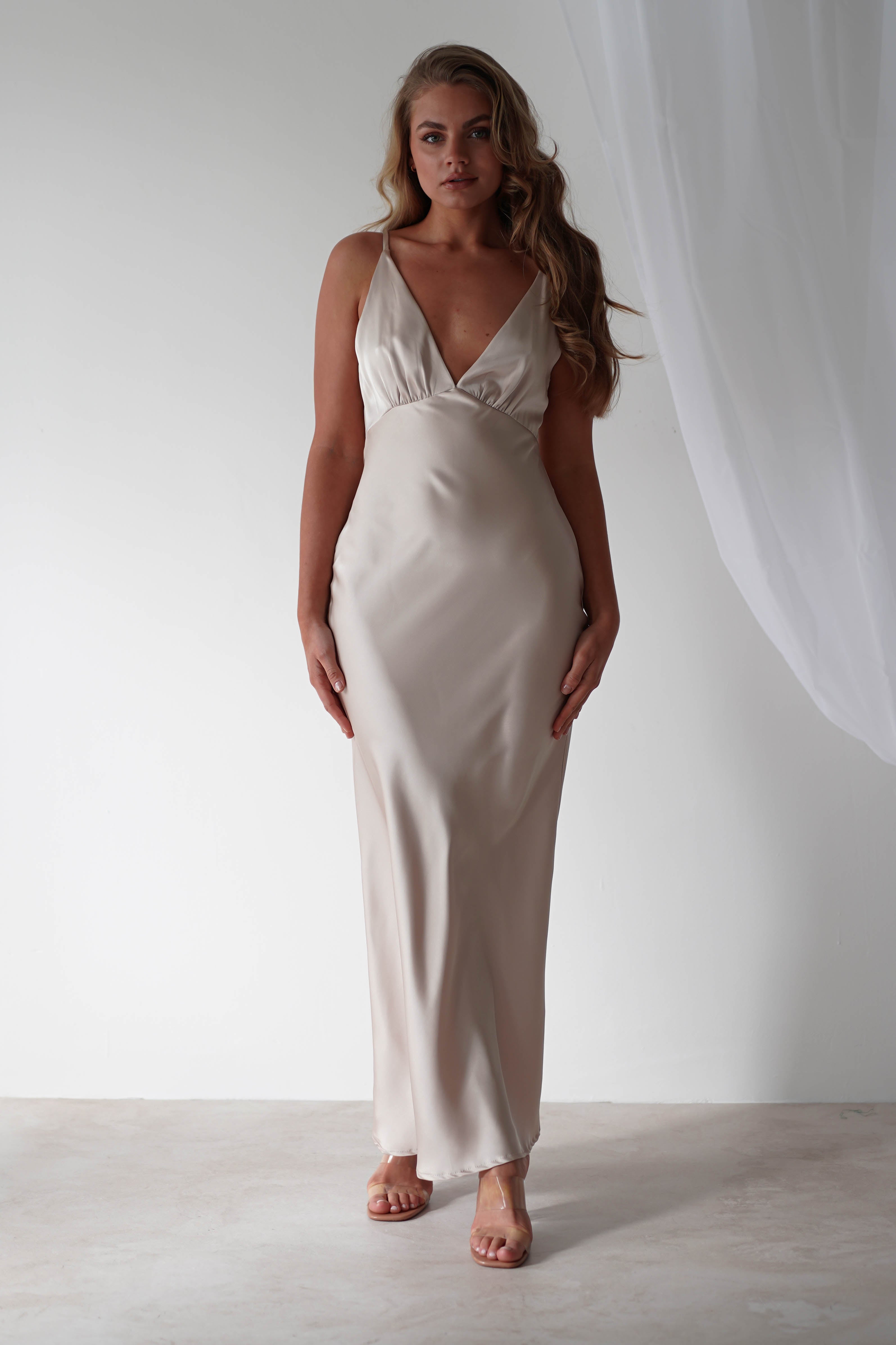 A woman in a sleek, light beige satin maxi dress stands on a light-colored floor. The dress, perfect as a wedding guest dress, features a deep V-neckline and is sleeveless. Her long hair flows over her shoulders as she stands against a simple white backdrop with a sheer curtain.