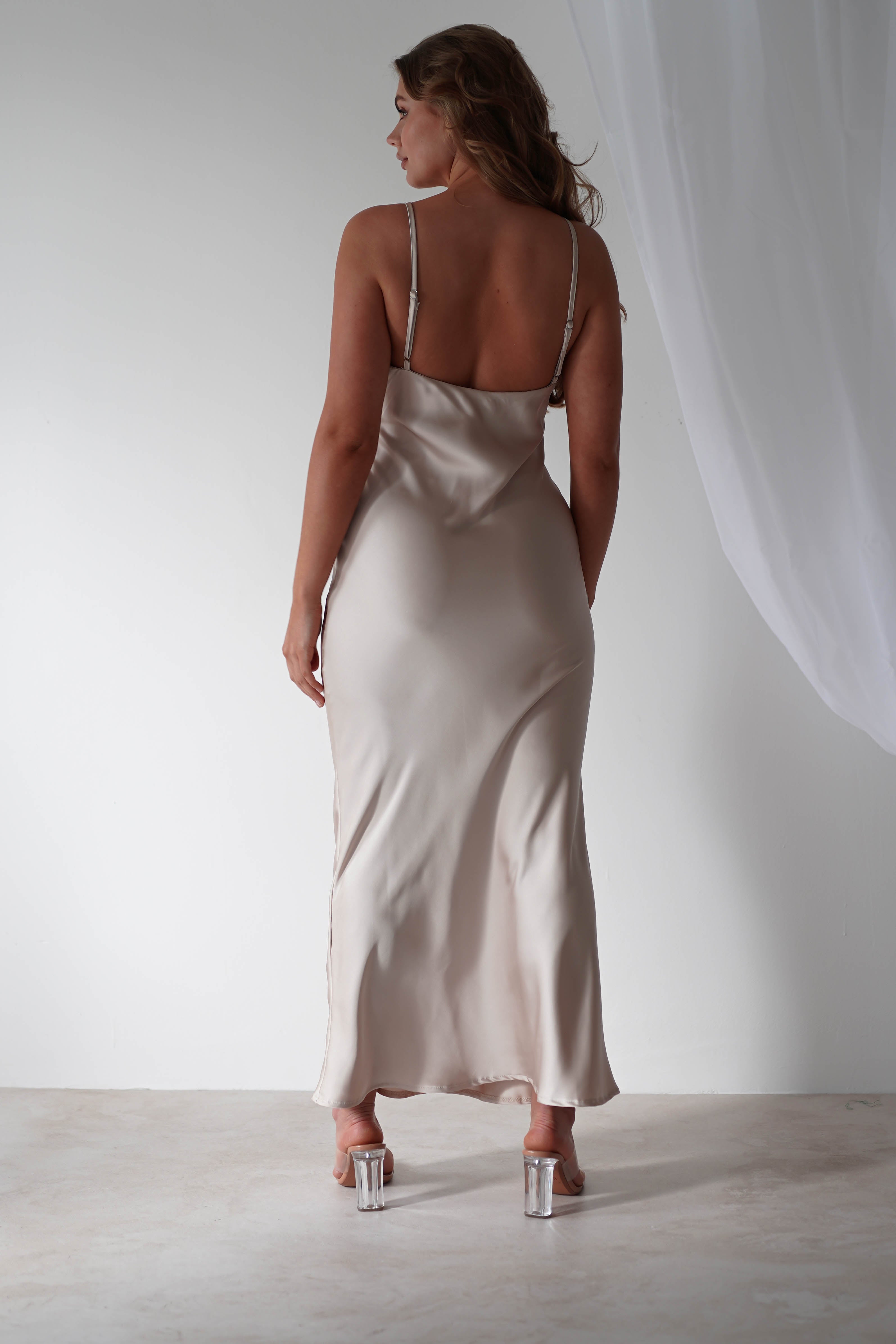 A woman stands with her back to the camera, wearing a long satin maxi dress with thin straps. Her hair is loose, and she is wearing high-heeled shoes. A white curtain drapes softly in the background against a plain wall.