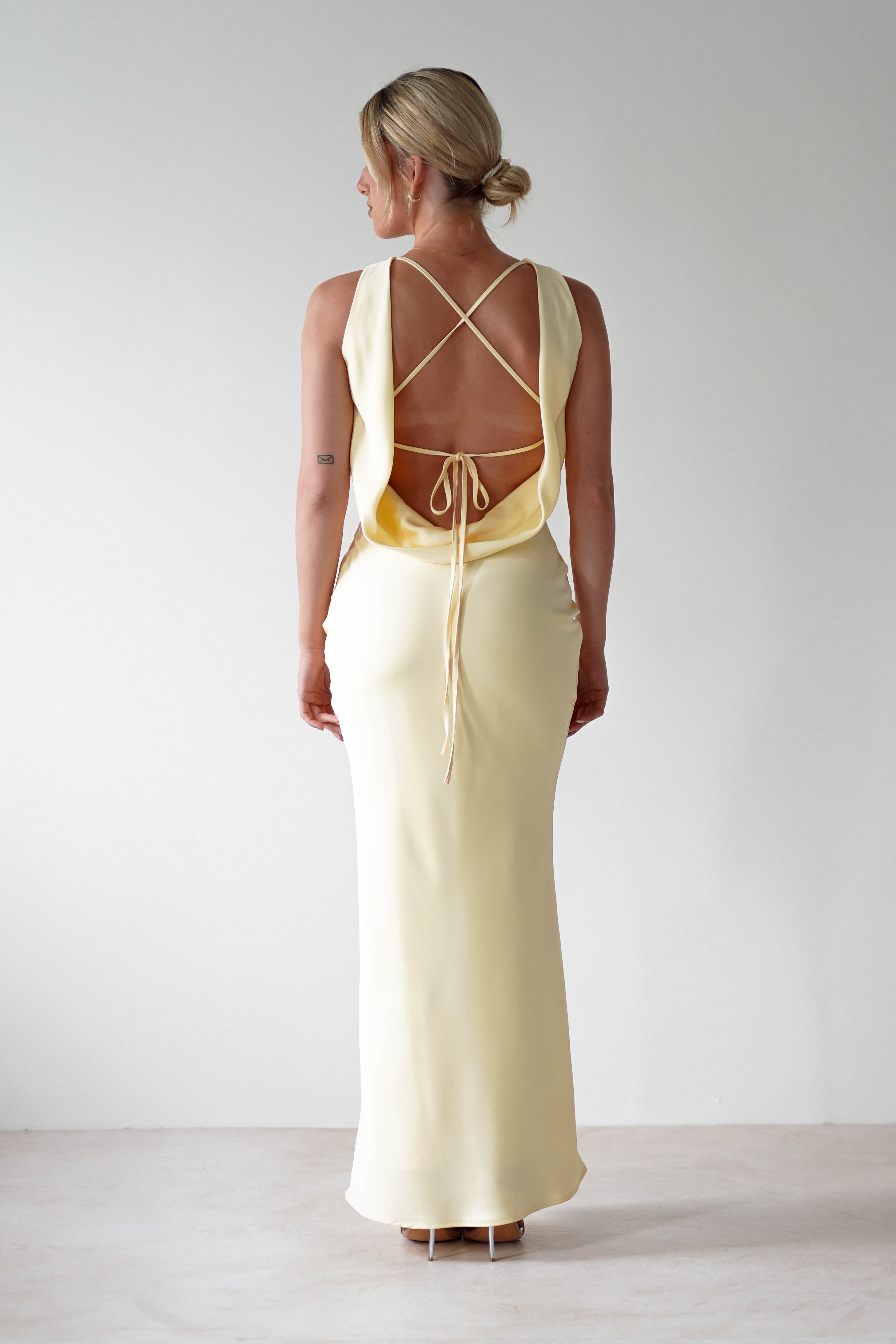 Rear view of a woman in a sleeveless, pale yellow satin maxi dress with an open back featuring crisscrossed straps and a tied detail. She has blonde hair in a low bun, perfectly capturing the elegance ideal for bridesmaids, against a plain white background.