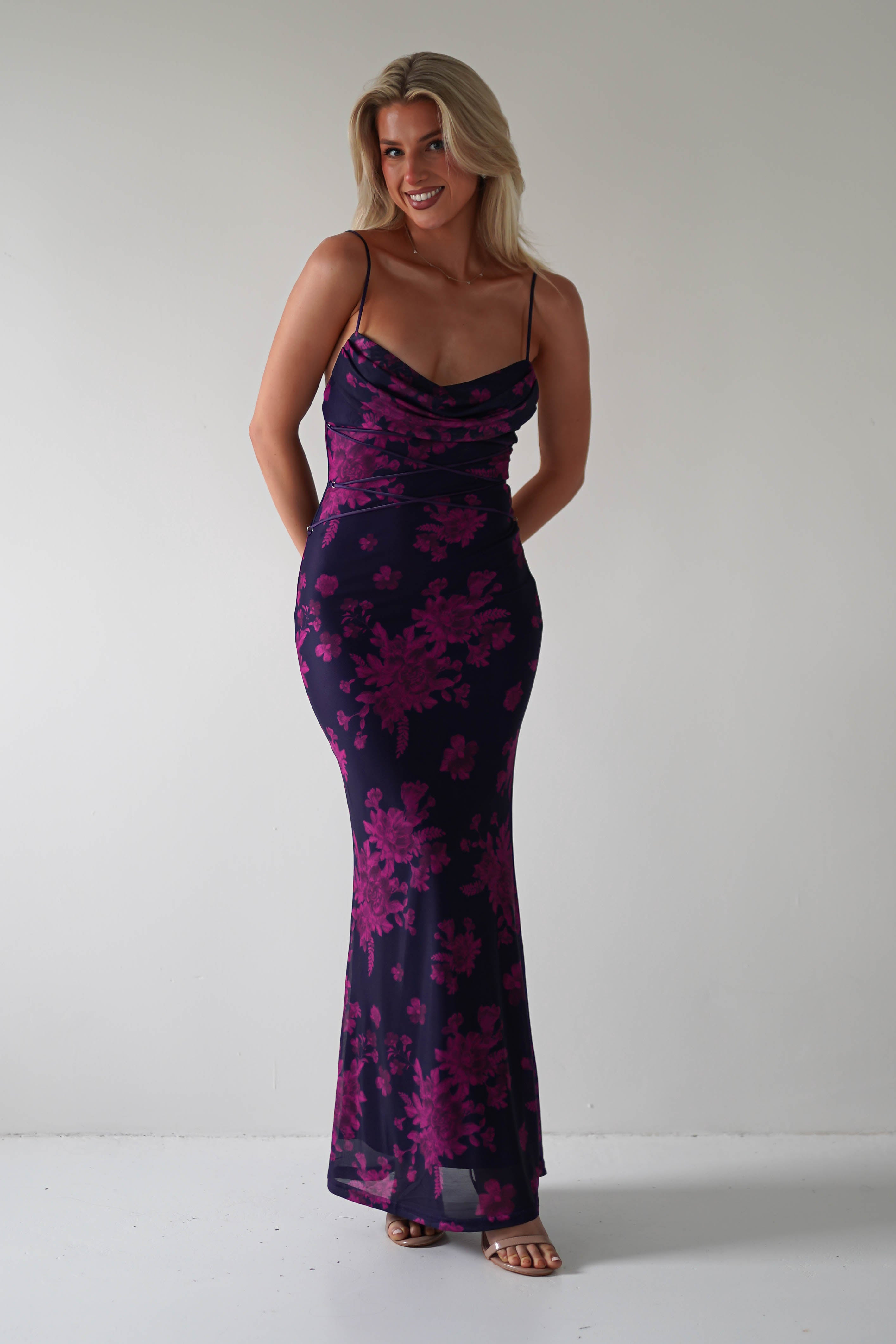 Person wearing a stretchy printed maxi dress with pink floral patterns, its floor-length design perfect for special occasions, and smiling against a plain light background.