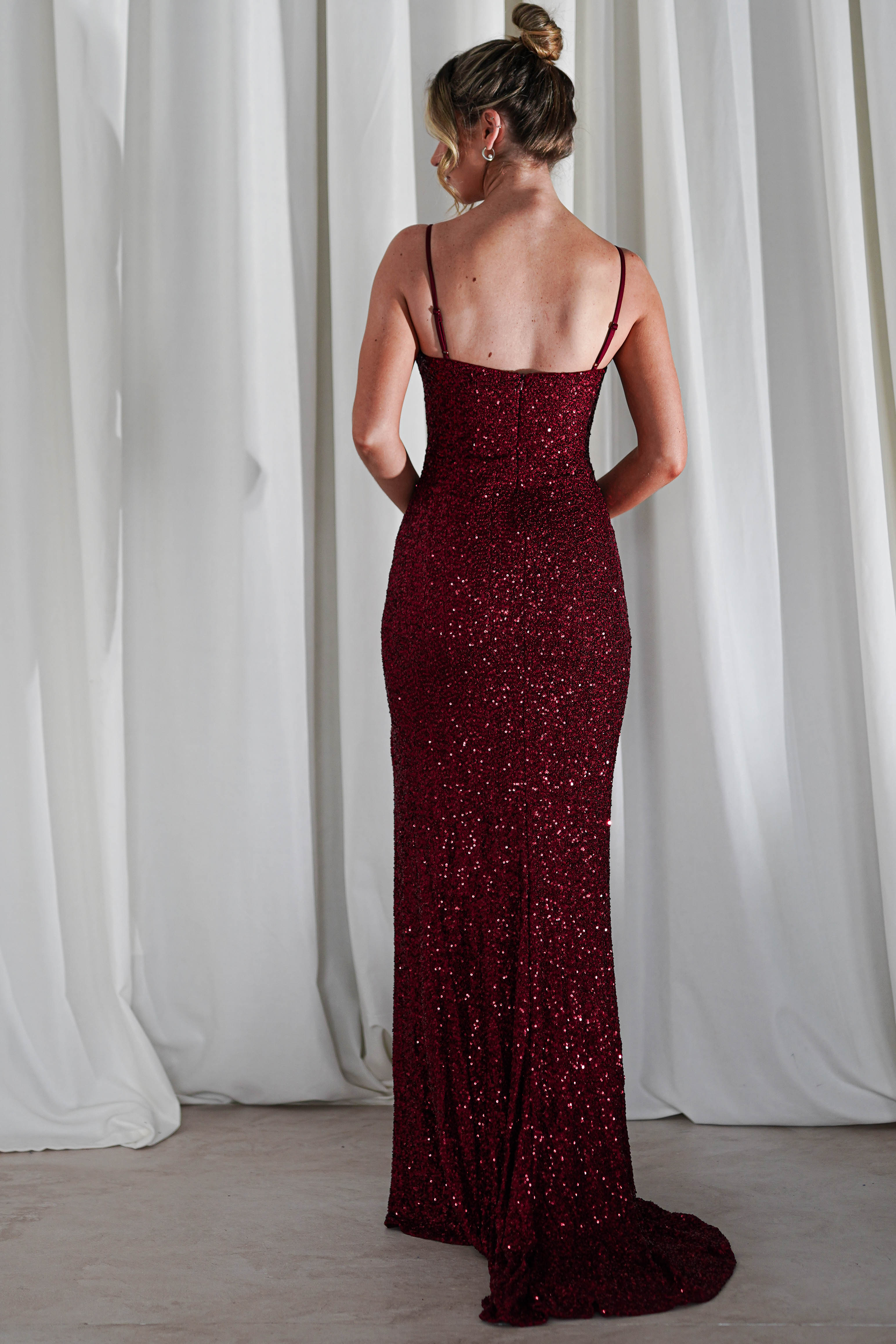 A woman in a floor-length, deep red sequin gown with a cowl neckline and thin straps stands with her back to the camera, her hair in a bun, in front of white curtains—perfect for formal events.