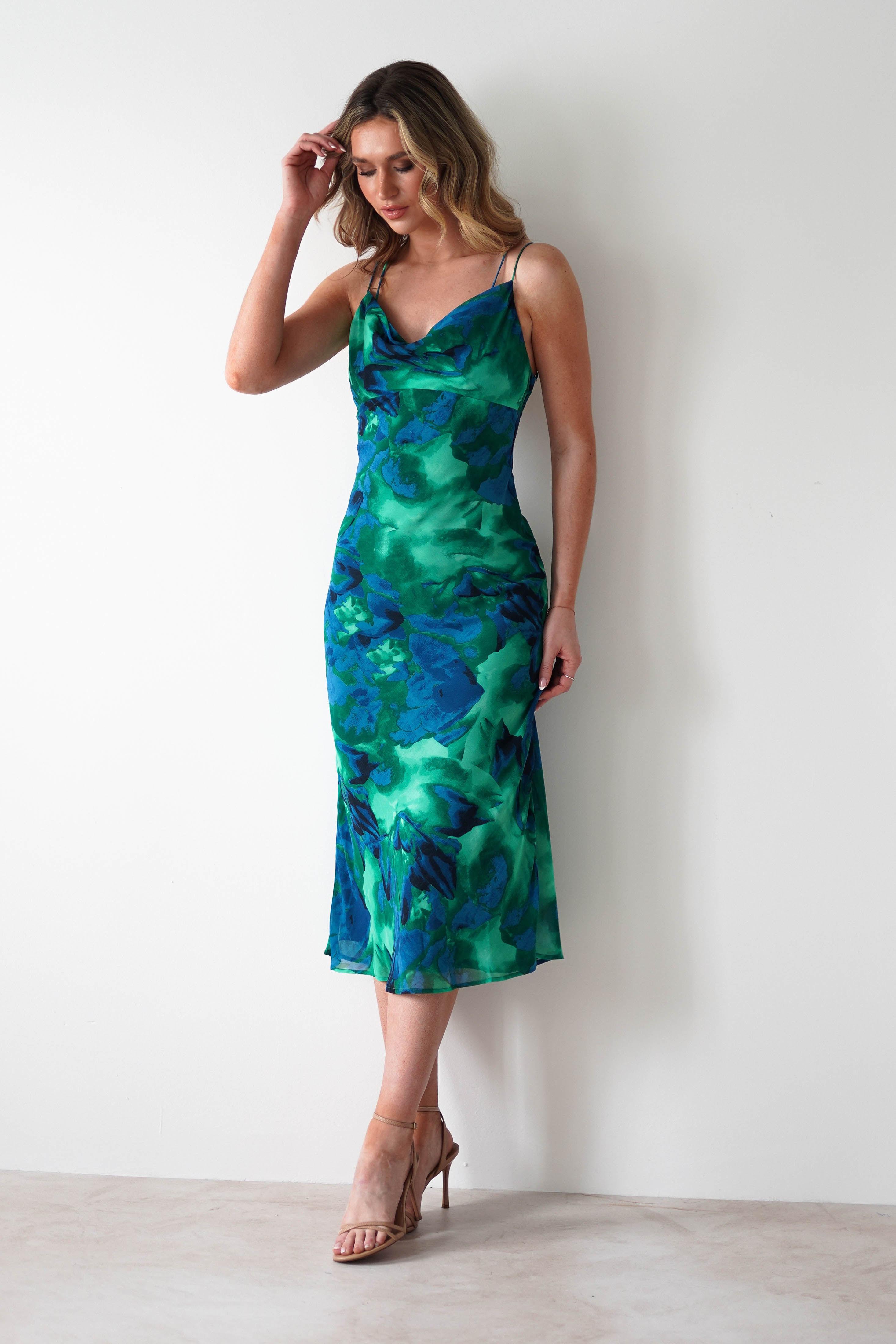 A woman stands against a white wall, wearing a sleeveless, green and blue printed midi chiffon dress with spaghetti straps and nude high-heeled sandals. Perfect as a wedding guest dress, she looks down while touching her hair.