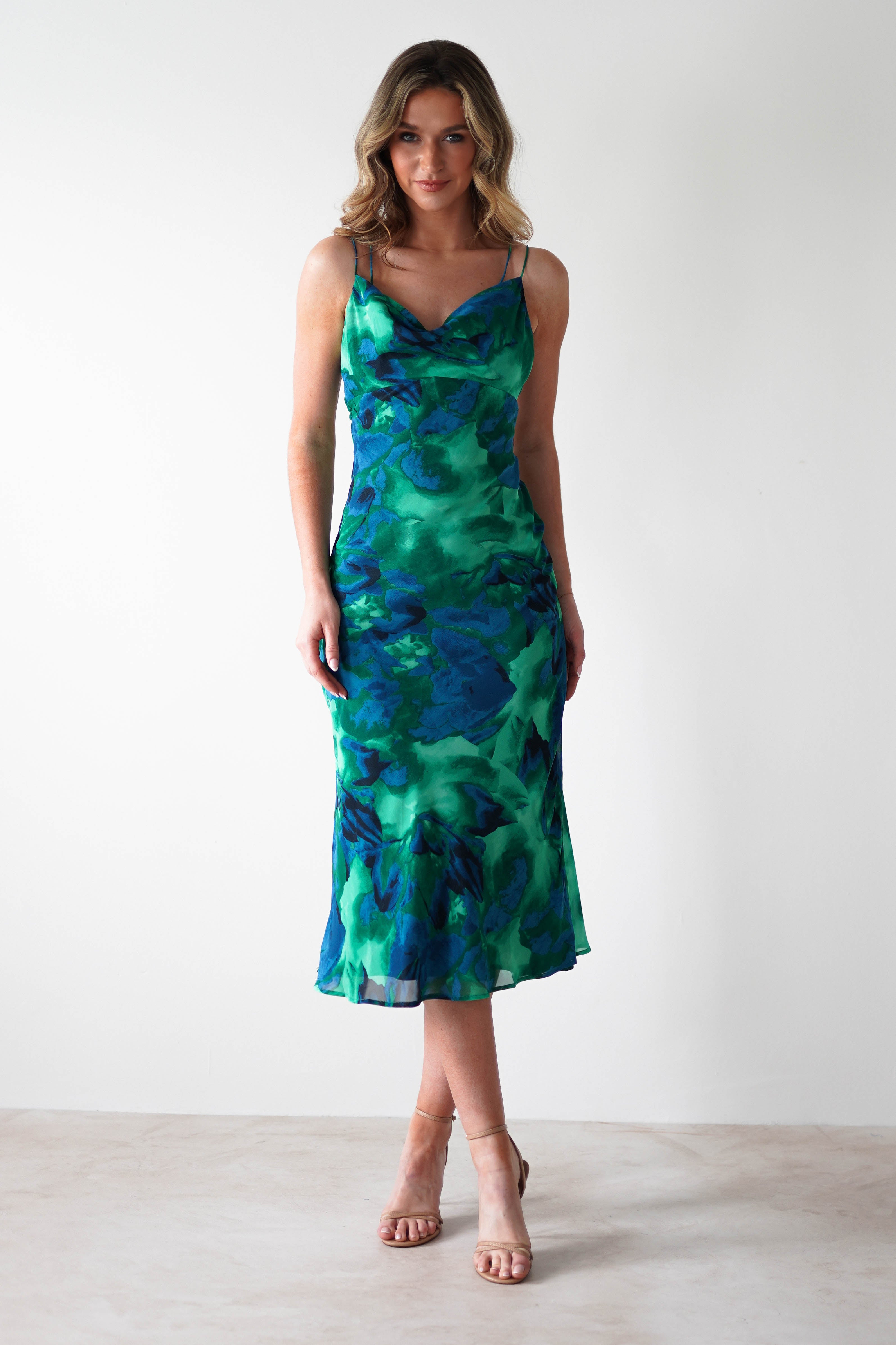 A woman stands against a white background, wearing a sleeveless green and blue Printed Midi Chiffon dress with thin straps and nude heels—an elegant choice for a wedding guest. She has wavy, shoulder-length hair and a slight smile.
