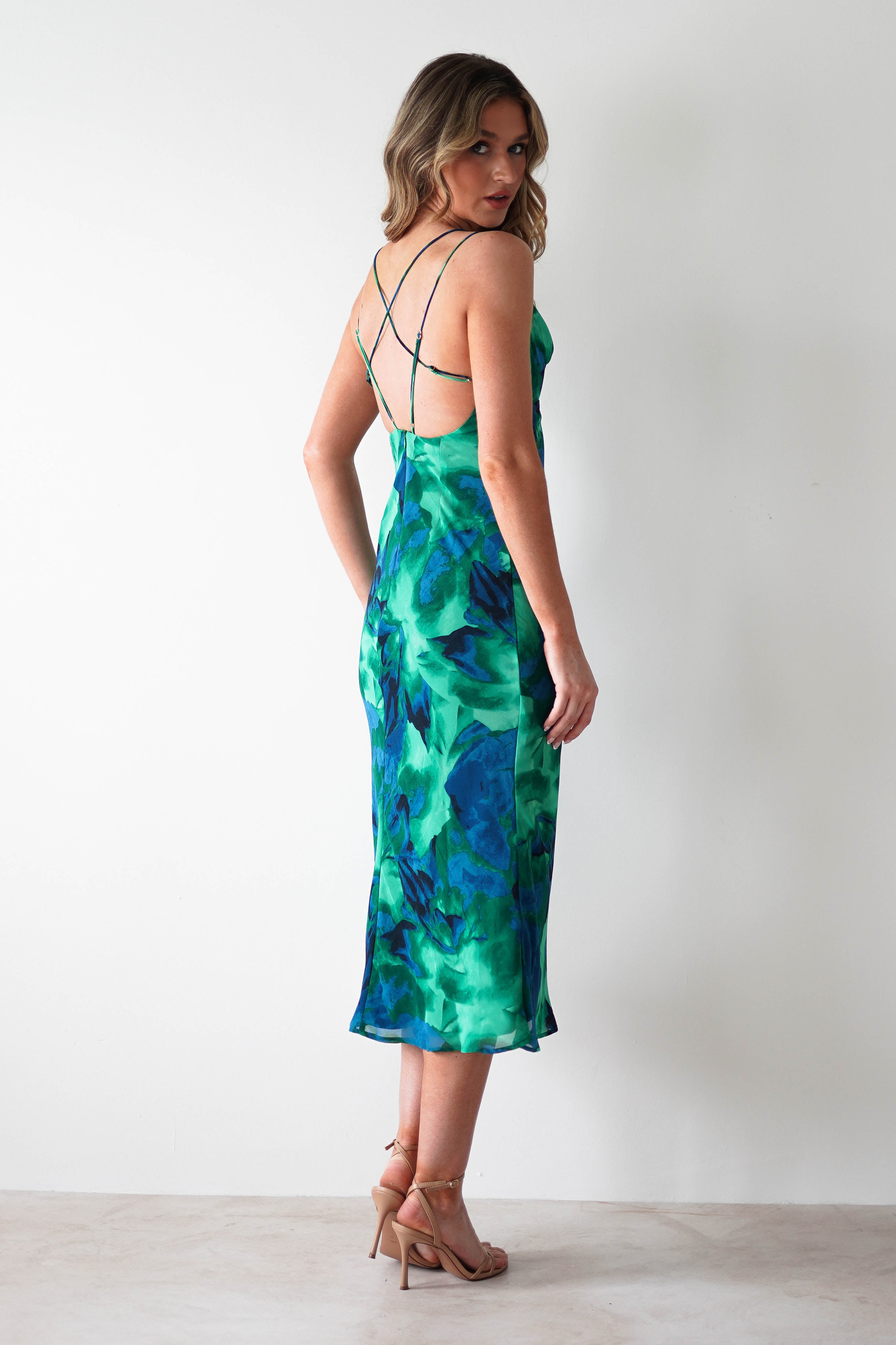 A woman wearing a green and blue printed midi chiffon, strappy back dress and nude heels stands against a plain white wall, looking over her shoulder—perfect for occasions or as a stylish wedding guest dress.