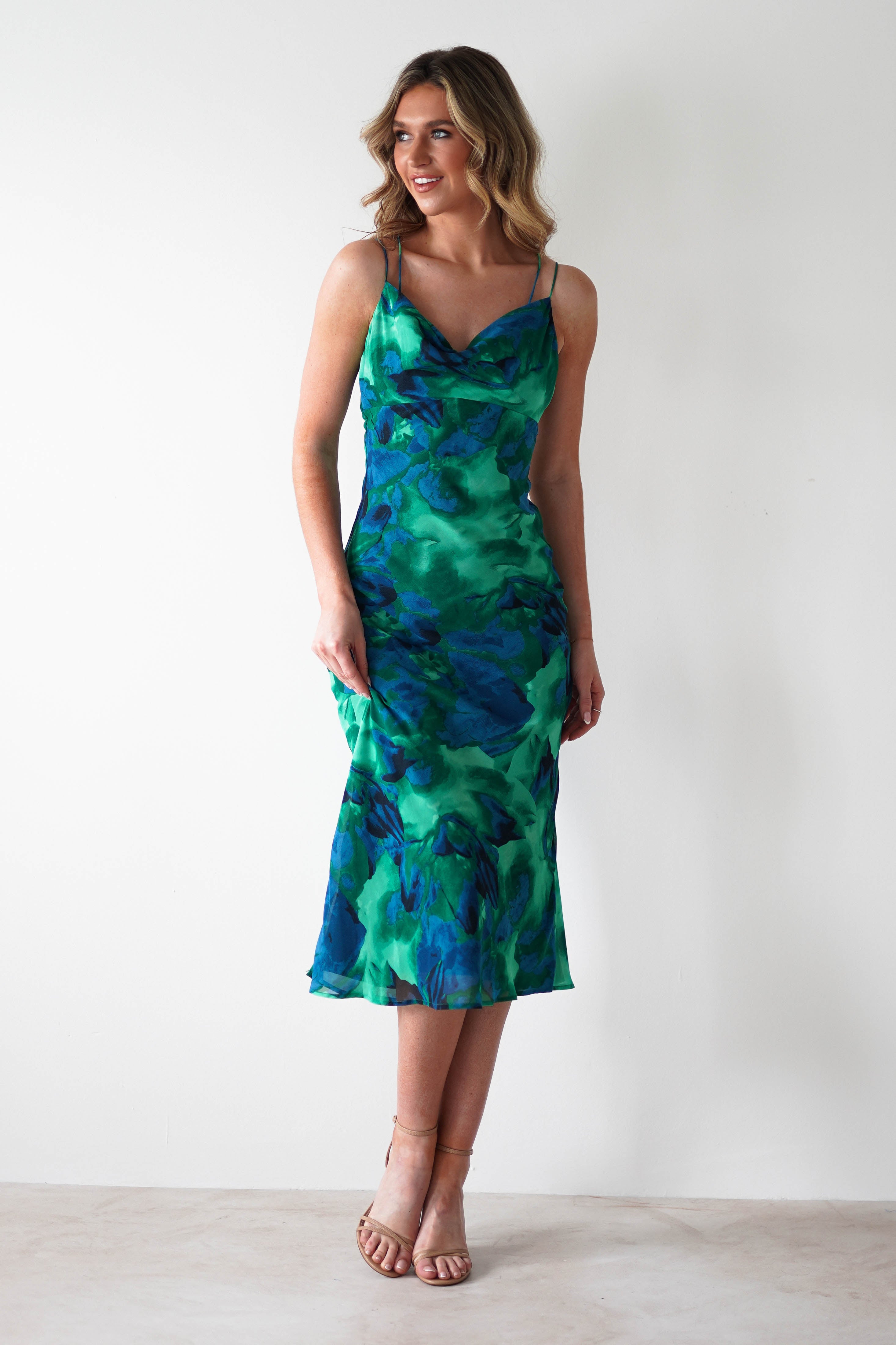 A woman stands against a plain white wall, wearing a printed midi chiffon dress with a green and blue abstract pattern—perfect for occasions as a chic wedding guest dress. She has wavy blonde hair and strappy nude heels.